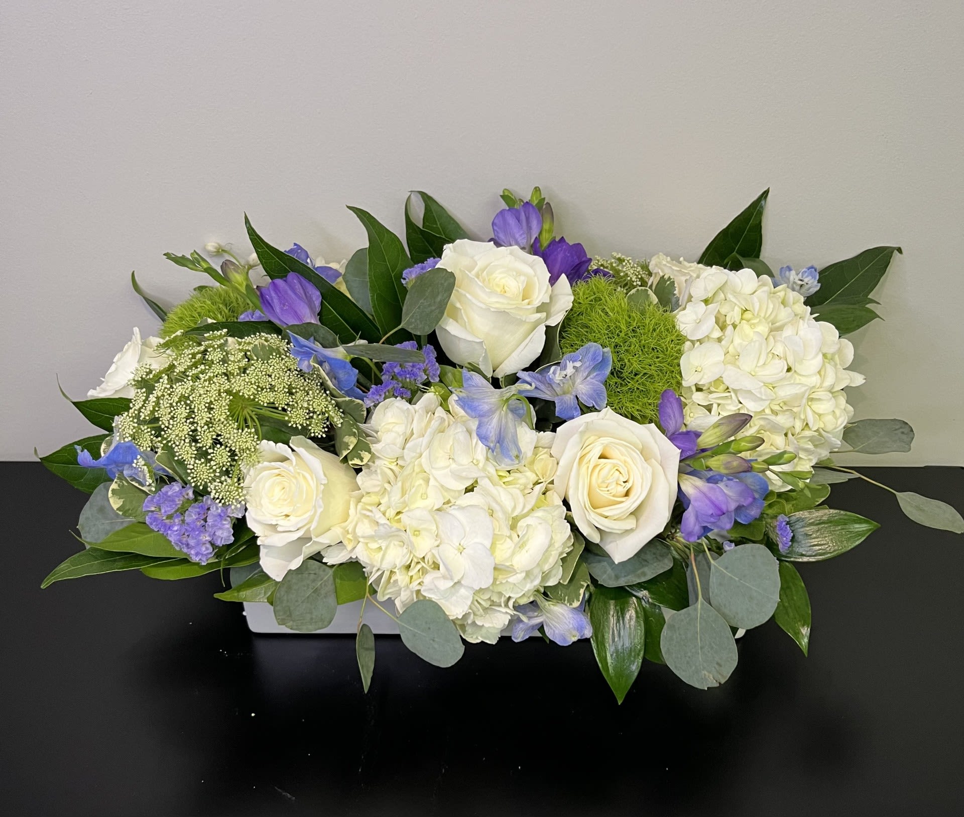 Blue Skies by Chasing Petals - A long and low white vase of beautiful white and green flowers with delicate pops of blue and lavender reminiscent of beautiful blue skies.