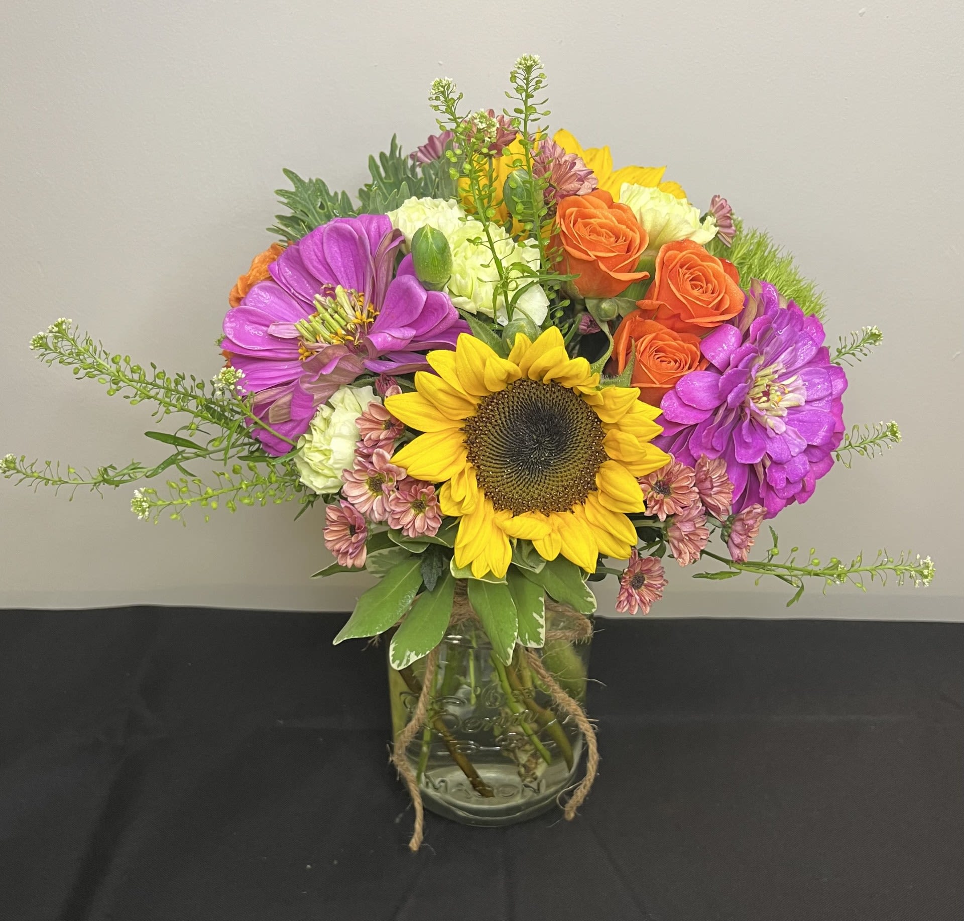 Sunkissed Fields by Chasing Petals - Brighten any room with the cheerful charm of our Sunkissed Fields arrangement. This vibrant bouquet features a radiant sunflower at its heart, surrounded by vivid orange roses, purple zinnias, and peach mums. Accented with fresh greenery and rustic twine, it's beautifully presented in a mason jar for a touch of country elegance. Perfect for birthdays, celebrations, or simply bringing a smile to someone’s day.