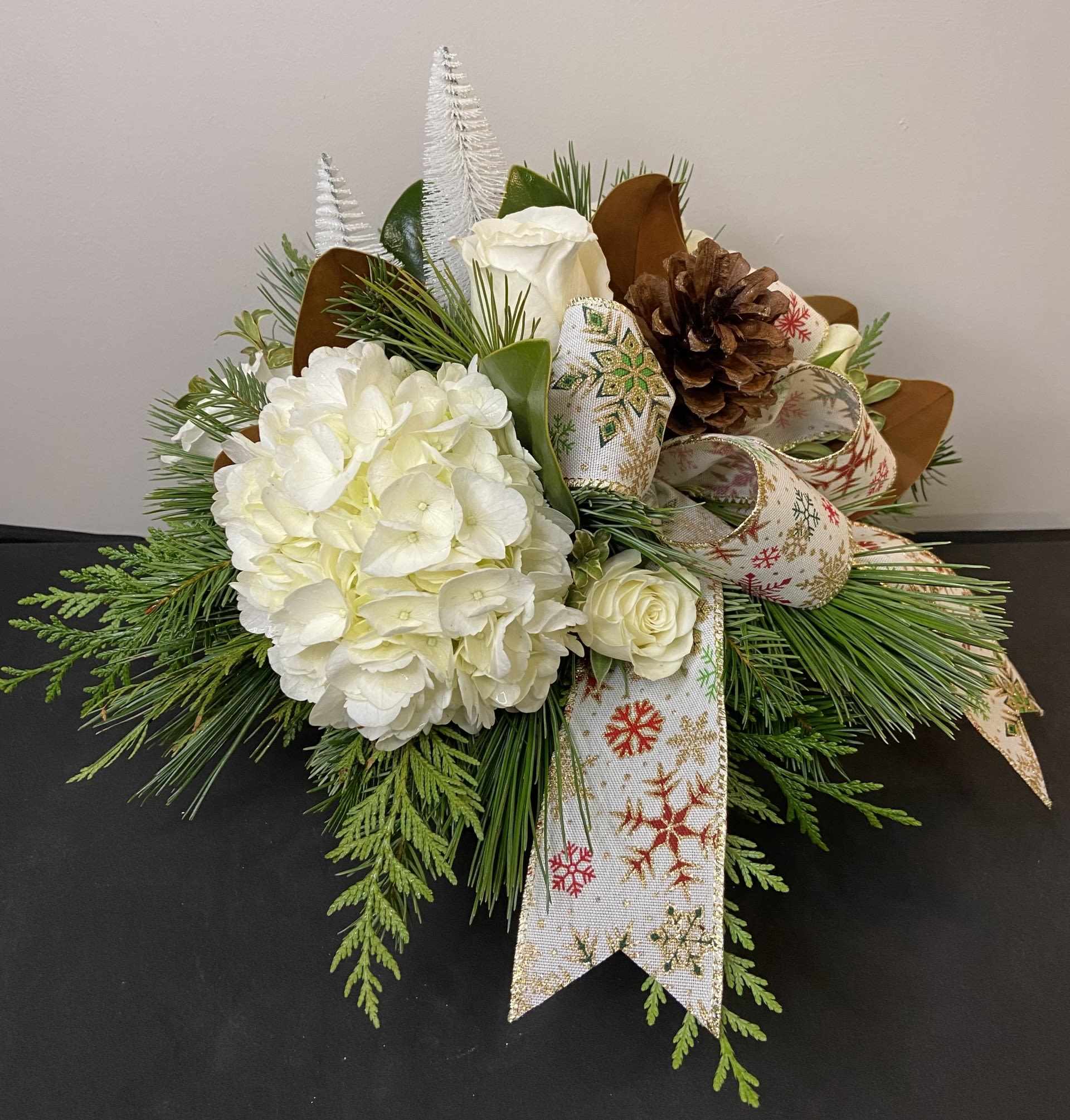 Snowfall Serenity by Chasing Petals - Our “Snowfall Serenity” arrangement brings the peaceful beauty of winter indoors. Soft white hydrangea and roses are nestled among lush evergreens, accented with pinecones, sleek magnolia leaves, and glittering snow-kissed elements. Finished with a festive snowflake ribbon, this arrangement adds a touch of cozy elegance to any holiday table or winter décor. Perfect as a centerpiece or seasonal gift. 
