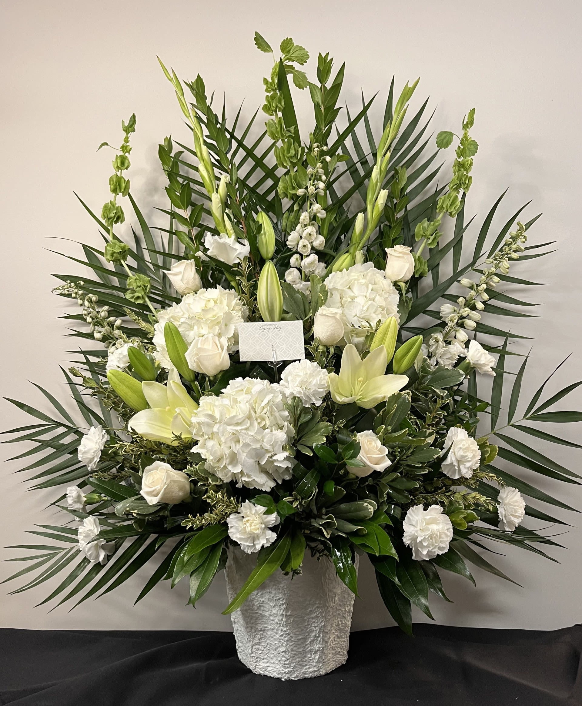 Tranquil Tribute by Chasing Petals - Tranquil Tribute is a heartfelt expression of sympathy, thoughtfully designed to offer comfort, peace and quiet reflection during difficult moments. Featuring soft white blooms gathered in lush, sculptural greenery, this elegant arrangement conveys grace remembrance and enduring love. Tranquil Tribute is a gentle and meaningful way to honour a life, share condolences, and bring a sense of calm to those who need it most.   Please note: While we always strive to include open lilies, we cannot guarantee they're availability. If substitutions are necessary, we will notify you at the time of your order. 