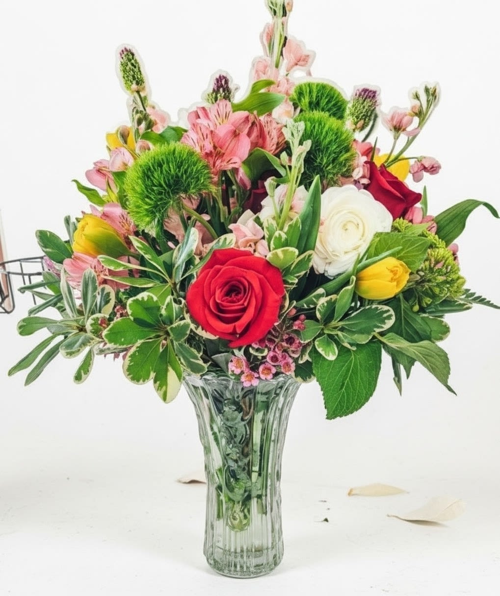 Mixed bouquet of red roses, yellow tulips, white blooms, and pink flowers in a clear glass vase