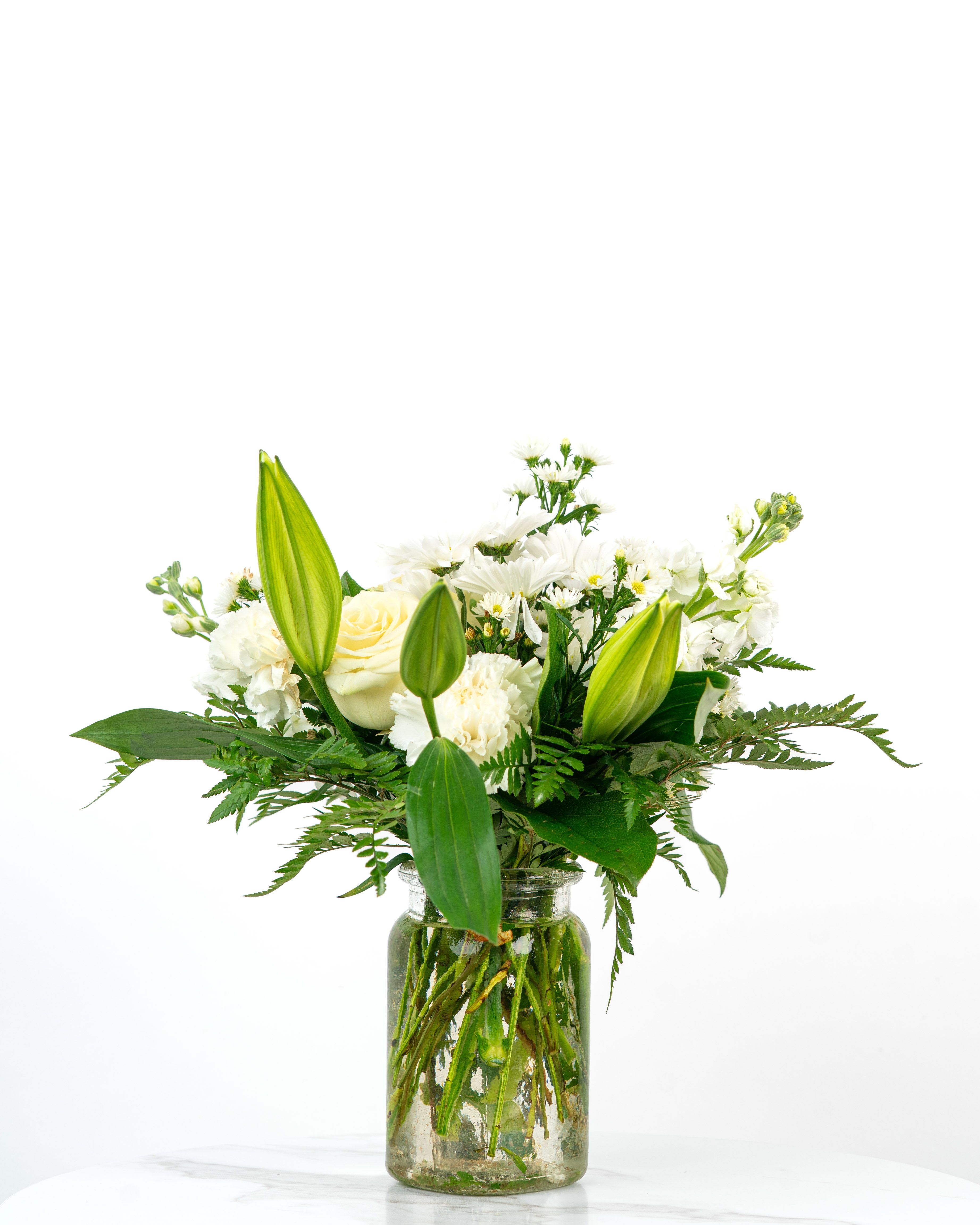 White lilies, roses, and other white flowers arranged in a clear glass jar vase