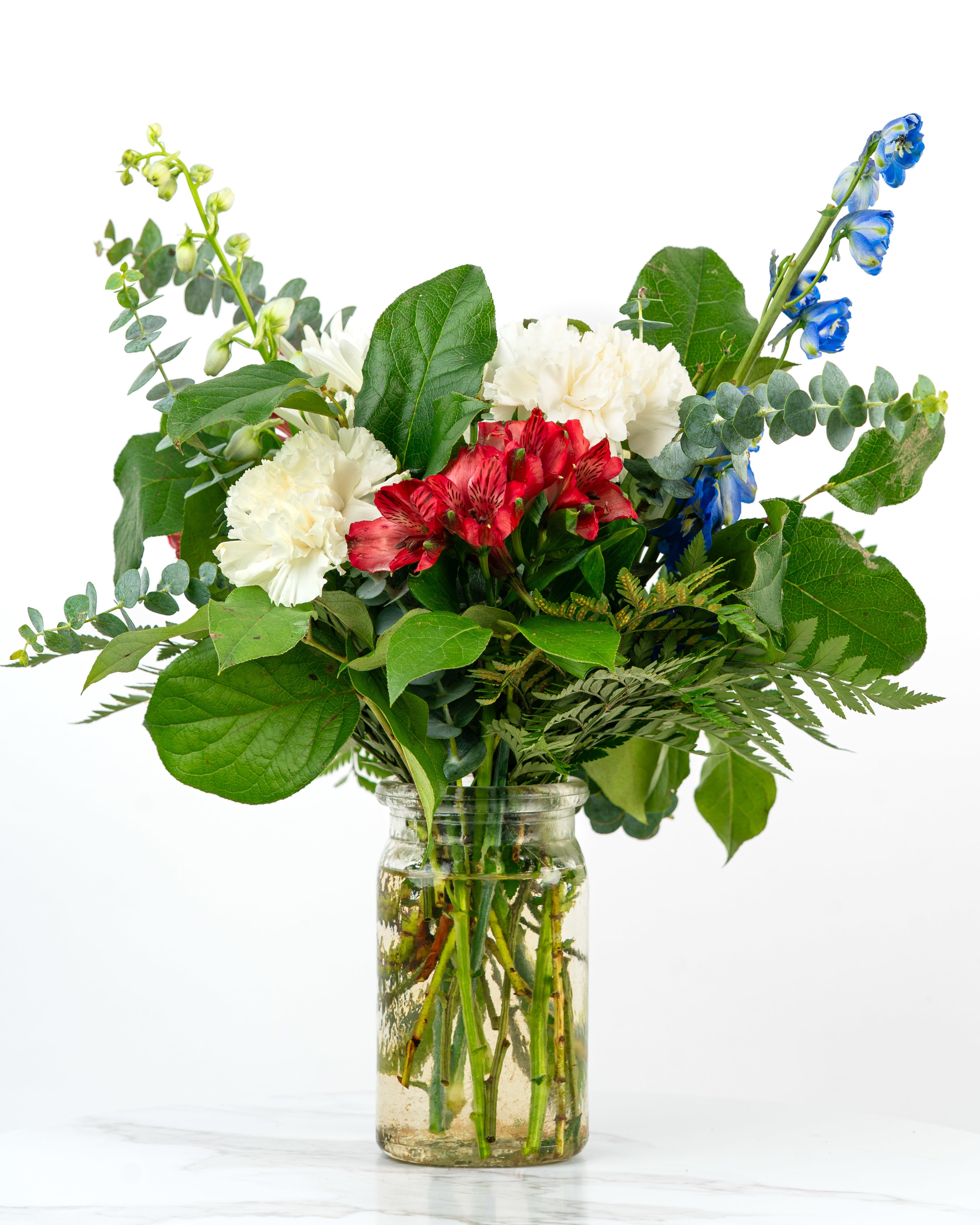 Red, white, and blue mixed flower arrangement with lush foliage in a clear glass jar vase