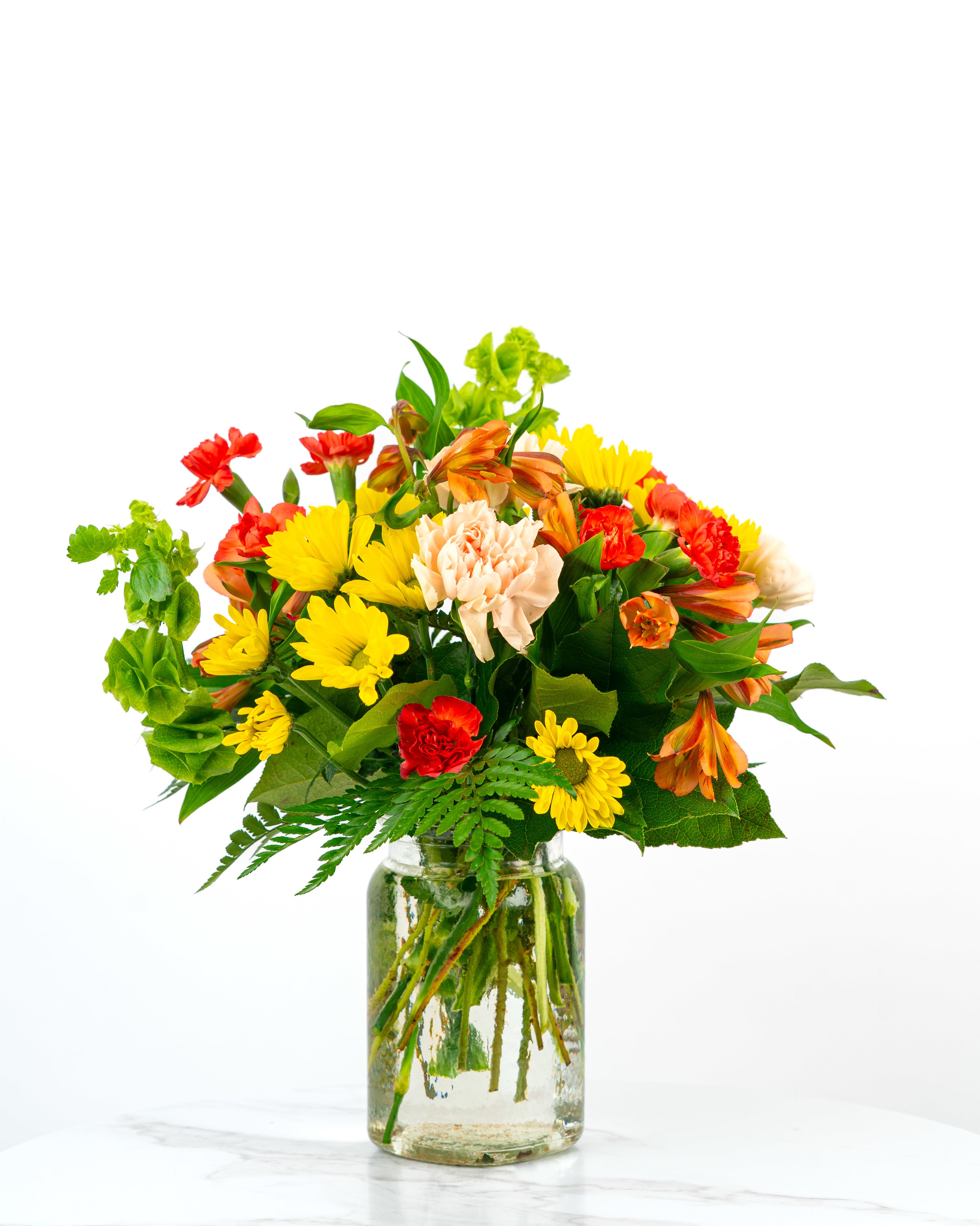 Bright mixed bouquet of yellow daisies, orange alstroemeria, and red and peach carnations in a clear glass jar.