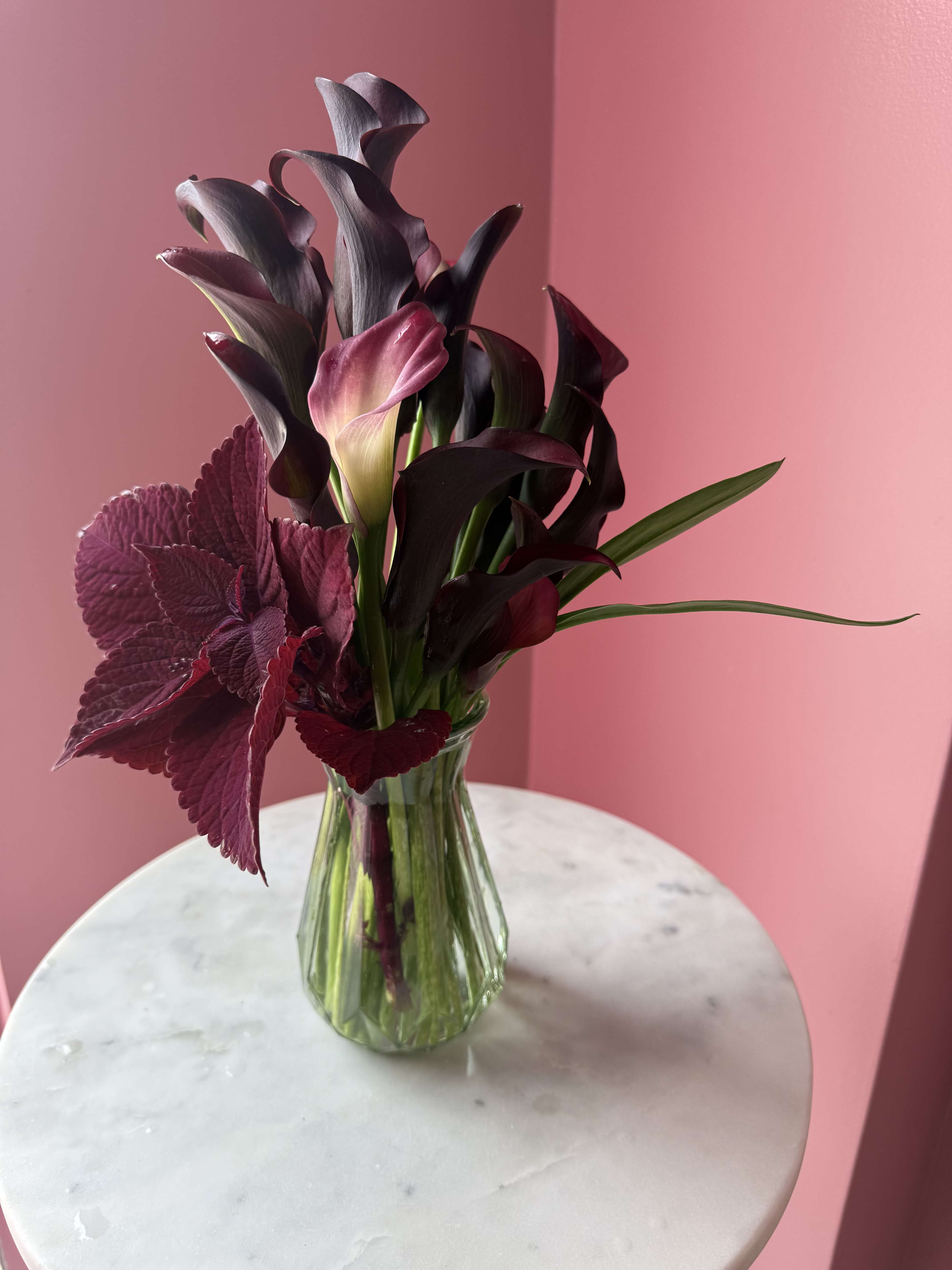 Velvet Noir  - Dark purple/plum  calla lilies with a pink-and-cream accent, burgundy foliage in a clear ribbed glass vase.
