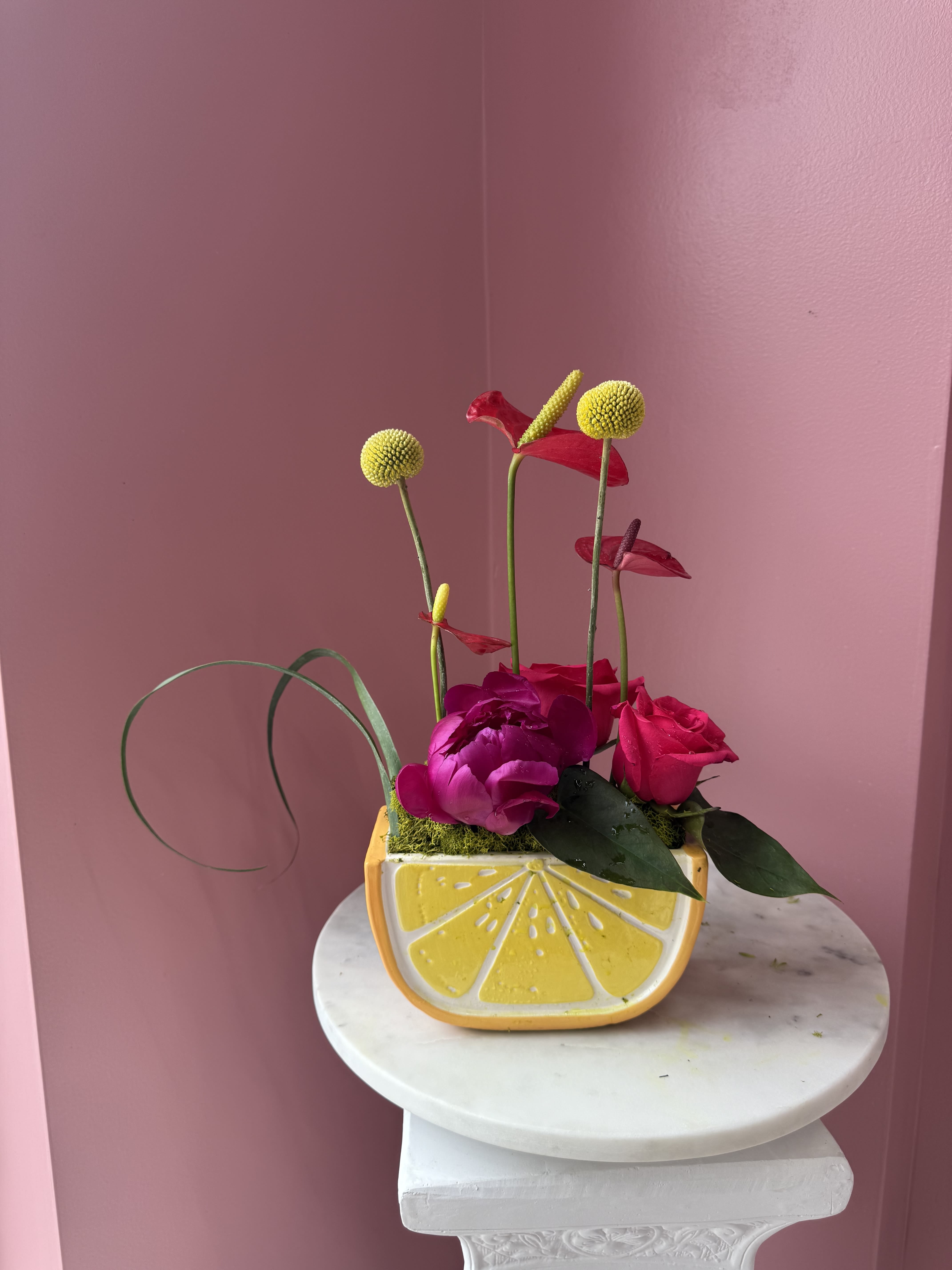 Main Squeeze  - Bright pink and magenta roses cluster low in a cheerful lemon-shaped ceramic container, while tall yellow billy stems and a few red anthurium-like blooms rise above for a playful, sculptural profile-perfect as a birthday bouquet, housewarming gift, or a just-because surprise. The vivid pinks and sunny yellow paired with touches of purple and green create an upbeat, modern look that will make the person you're thinking of feel noticed and delighted.  Hand‑arranged with glossy petals set against decorative moss, this compact arrangement balances smooth ceramic, soft floral texture, and a whimsical silhouette that fits on a kitchen counter or entry table. The low base keeps blooms close together for a full, lush appearance while the taller stems add height and movement; ideal for brightening a home or office without taking up much space. Care: keep vase water fresh and trim stems every few days to prolong bloom life.