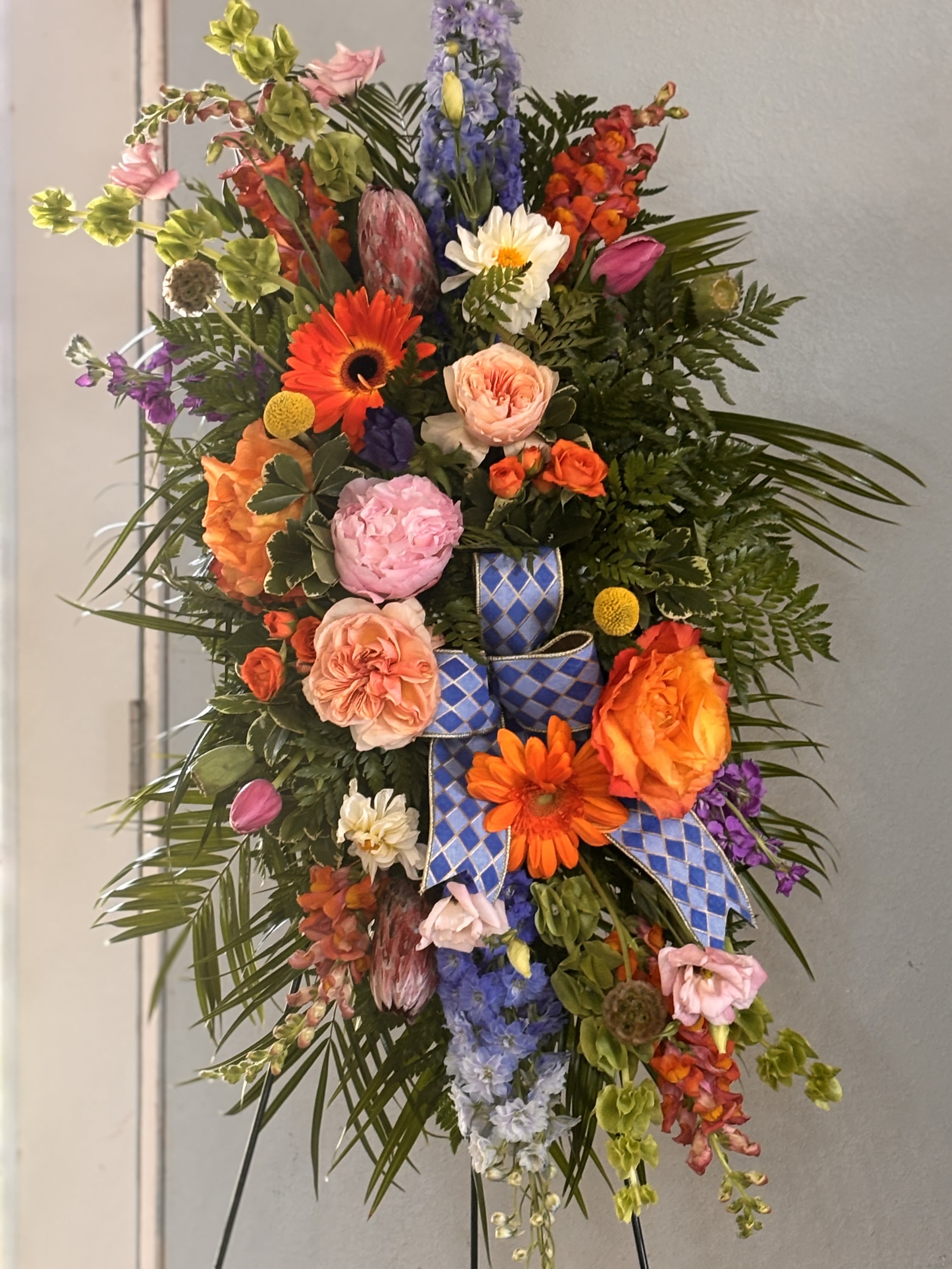 Bright mix easel  - A bold, tall standing floral spray that reads like a joyous, ceremonial statement-perfect as a celebration bouquet or a respectful memorial tribute. This standing floral arrangement features a bright mix of orange, pink, purple and blue blooms so the person you're honoring will feel both seen and celebrated; its vertical, asymmetrical silhouette gives immediate presence in any room or chapel.  The spray is densely layered with roses, large gerbera daisies, snapdragons, delphinium, lisianthus, protea, dendrobium orchids, chrysanthemums and other seasonal accents set over abundant dark greenery and palm fronds. Tall spikes and cascading elements add height and movement while blue diamond-pattern and blue plaid ribbon streamers weave through the composition for a crisp, ceremonial contrast. Mounted on a standing easel, this piece reads formal and festive at roughly traditional standing-spray scale.  Ideal for celebrating a life, marking a milestone, or sending a bold sympathy statement, this arrangement brings vibrant color and texture to the moment. Order with your preferred delivery date and our team will ensure the arrangement is securely mounted on the easel and arrives ready to display.