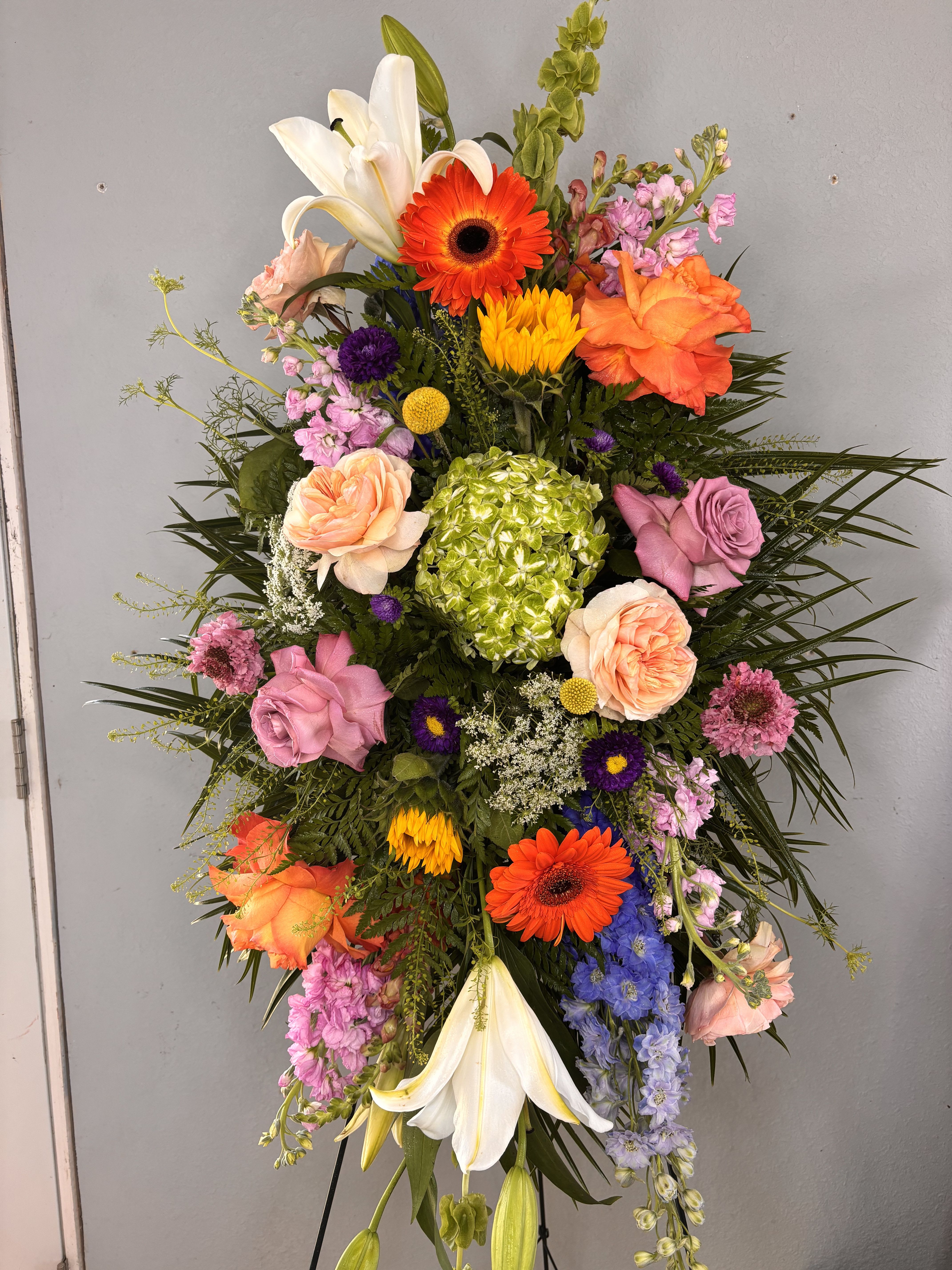 The Barbara standing easel  - Bright, tall, and unmistakably celebratory, this standing easel spray brings together lilies, roses, gerbera daisies, hydrangea and snapdragons in a mixed-color palette perfect for sympathy, memorial services, or a bold congratulatory display. The vertical, cascading shape reads formal and respectful on an easel yet lively in color-she'll see white lilies and peach-pink roses as comforting focal points while bright orange gerberas and purple accents lift the mood.  Built as a layered, oval-style standing arrangement on a metal easel with a pedestal vase option, the design features a central green hydrangea cluster, long trailing stems, airy filler and rich dark foliage for contrast. Expect a full, textured composition with strong vertical movement: large lilies, clustered roses and hydrangea, snapdragons for height, plus carnations, stock and daisy fillers to soften edges. The substantial presentation makes it suitable for funeral or memorial use, a public celebration, or an event centerpiece.  Choose this easel spray when you want the recipient to feel remembered and celebrated-it makes a thoughtful tribute at services or a striking statement for anniversaries and congratulations. Sizes and placement are event-style; stems are arranged for display on the included easel and in a pedestal vase when requested. For best longevity, keep the vase water fresh if displayed off the easel and away from direct heat.