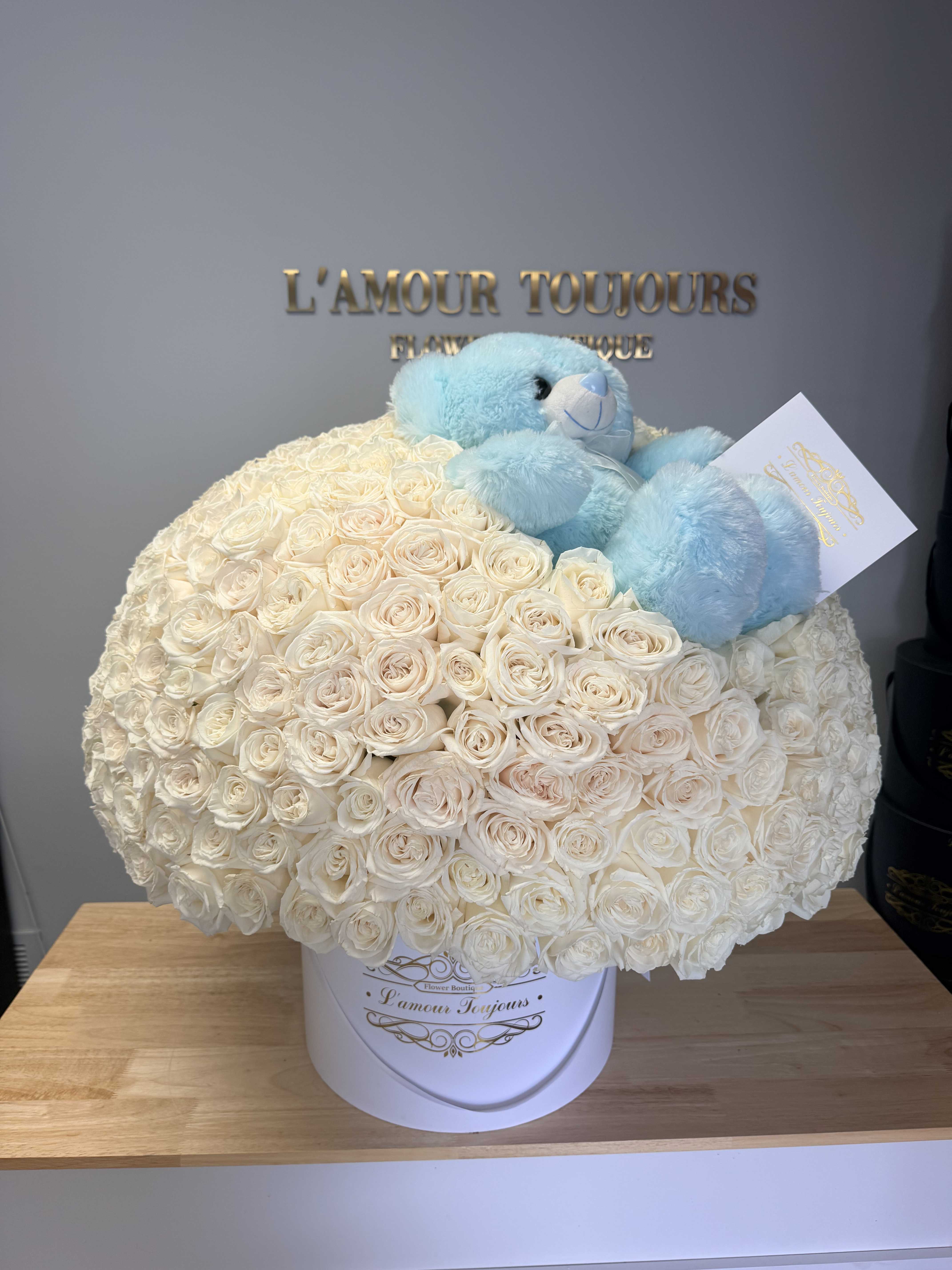 White rose arrangement in a round box with a blue teddy bear on top