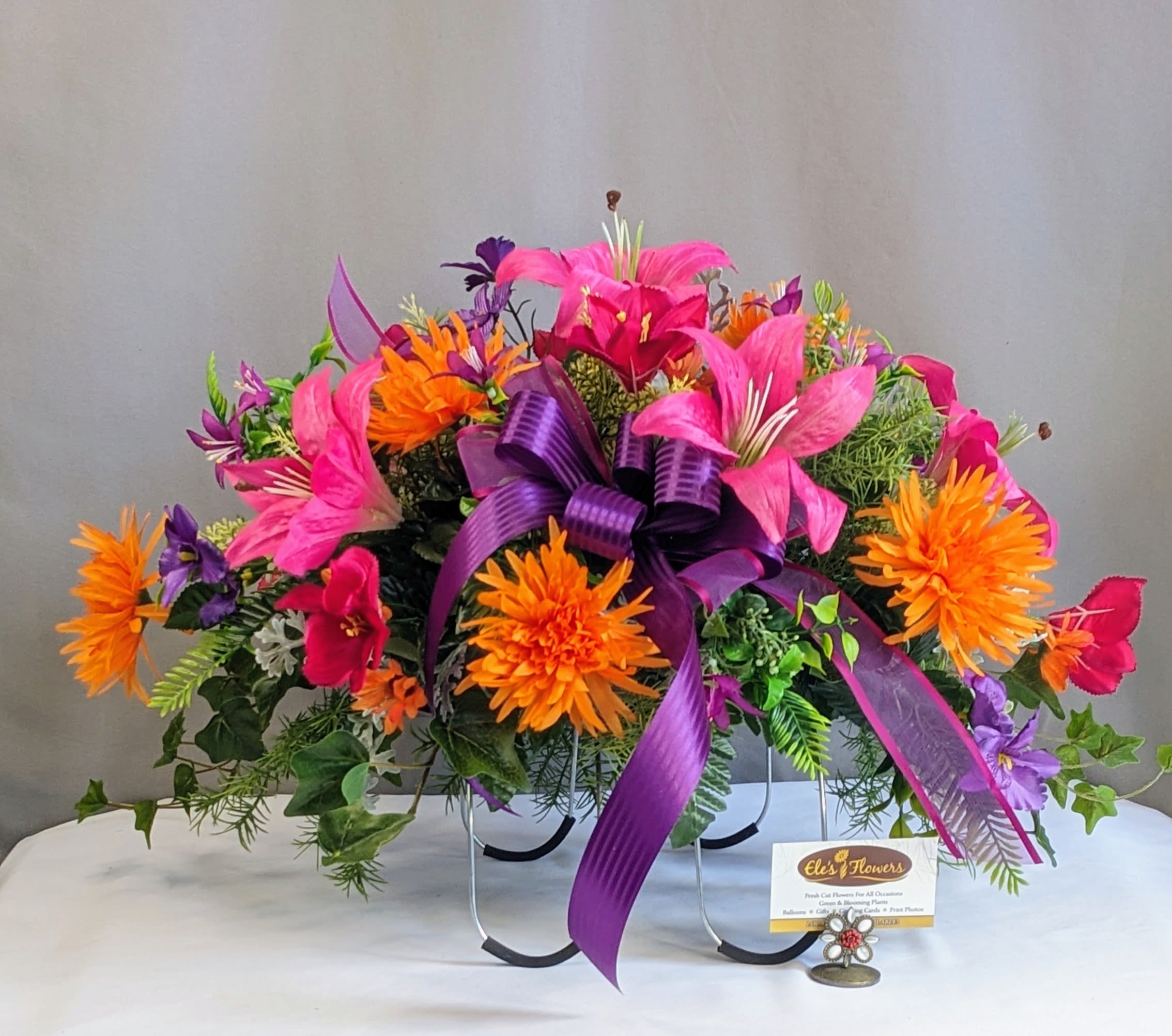Memorial Gravestone Topper - Artificial Arrangement - Honor the memory of a loved one with a beautifully crafted memorial silk arrangement designed especially for gravestone toppers. Featuring a heartfelt blend of rich pinks, purples, and vibrant oranges accented with a flowing purple ribbon, this arrangement brings warmth, beauty, and remembrance to any memorial setting. Thoughtfully created using high-end artificial flowers, it offers a lifelike appearance while remaining weatherproof for lasting outdoor display throughout the seasons. Its adjustable design allows it to securely fit most gravestone sizes with ease. Perfect for Memorial Day or any time of remembrance, this tribute serves as a meaningful way to celebrate and honor those who are forever in our hearts.  We would be happy to deliver this to your loved one's gravestone, please provide information on location in the special instructions or give us a call at 715.644.3537  Approximate Dimensions: 16"H x 25"W