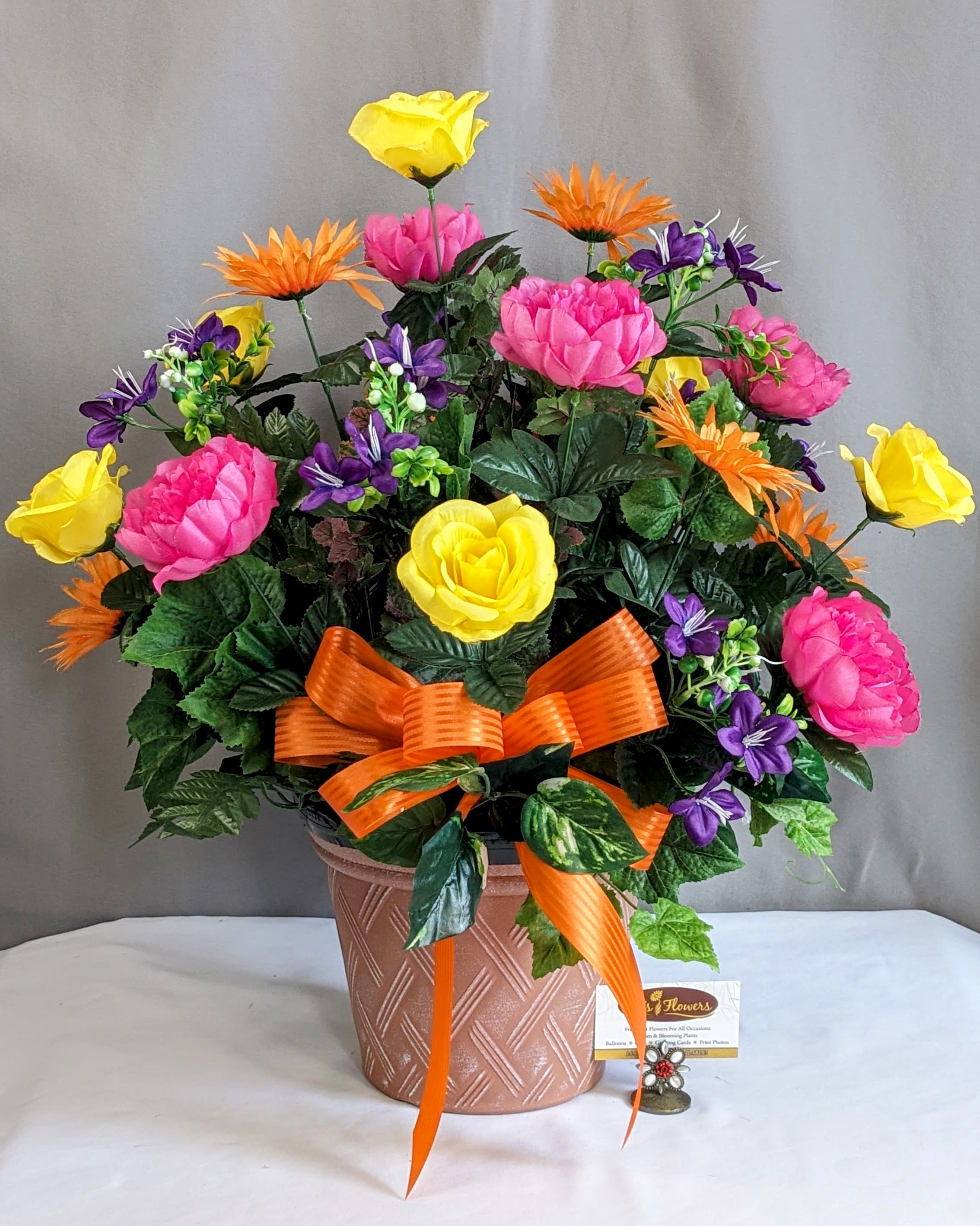 Memorial Planter Mixed Color - Artificial Arrangement - Honor and remember a cherished loved one with this beautifully crafted memorial silk arrangement, thoughtfully designed for placement at a graveside or cemetery memorial. Created in a decorative container designed to fit into most standard cemetery metal flower pot stands, this arrangement offers a meaningful and lasting tribute for Memorial Day or year-round remembrance. Featuring a vibrant blend of cheerful yellows, bright pinks, rich purples, and warm orange accents finished with an elegant coordinating ribbon, this design brings warmth, beauty, and comfort to any memorial setting.  Thoughtfully crafted using high-end artificial flowers and lush greenery, this weather-resistant arrangement is designed to maintain its beauty through the outdoor elements season after season. A heartfelt way to celebrate, honor, and remember those who remain forever in our hearts. Cemetery stands are not included.  We would be happy to deliver this to your loved one's gravestone, please provide information on location in the special instructions or give us a call at 715.644.3537  Approximate Dimensions: 25"H x 25"W