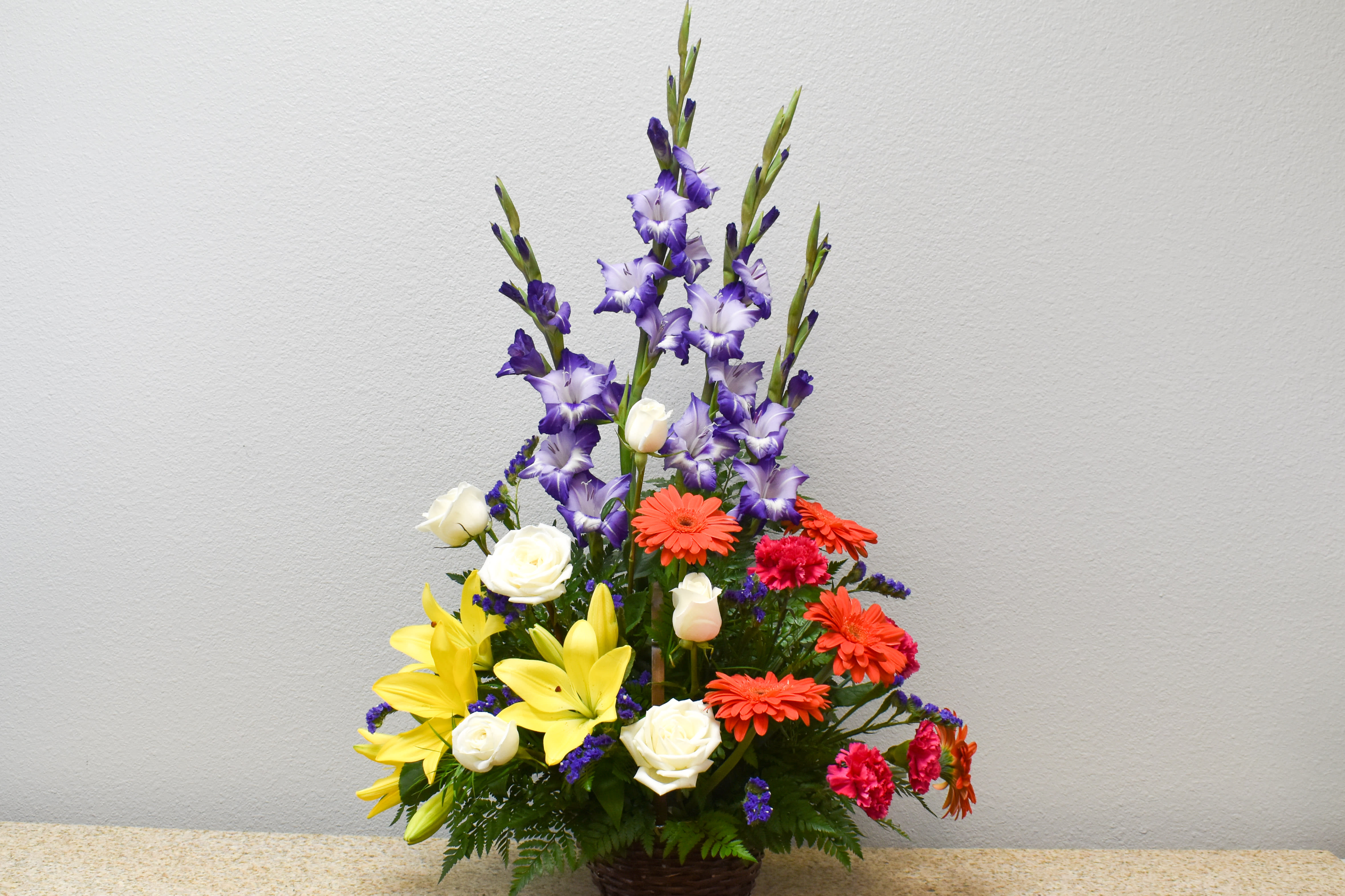 Tall mixed flower arrangement with purple spikes, yellow lilies, and orange daisies in a basket