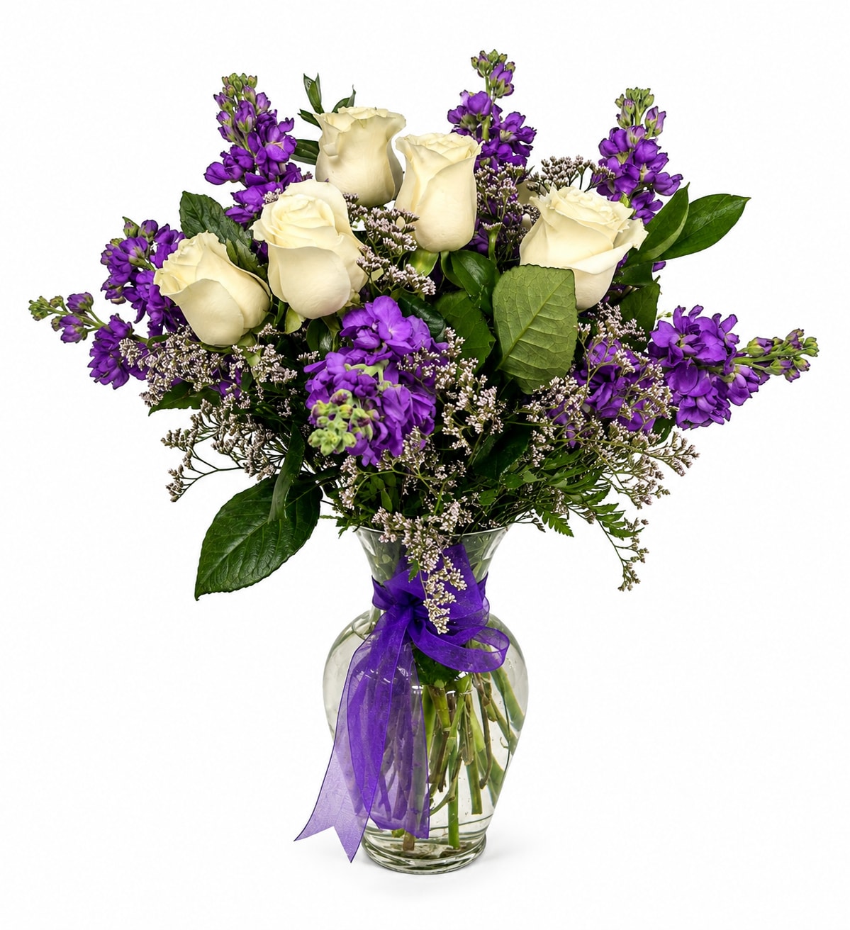 Enchantment  - A timeless arrangement of crisp white roses accented with vibrant purple stock and delicate filler flowers, beautifully arranged in a clear glass vase adorned with a purple ribbon. This classic design offers a graceful blend of elegance and charm, making it perfect for birthdays, sympathy, anniversaries, or any special occasion.