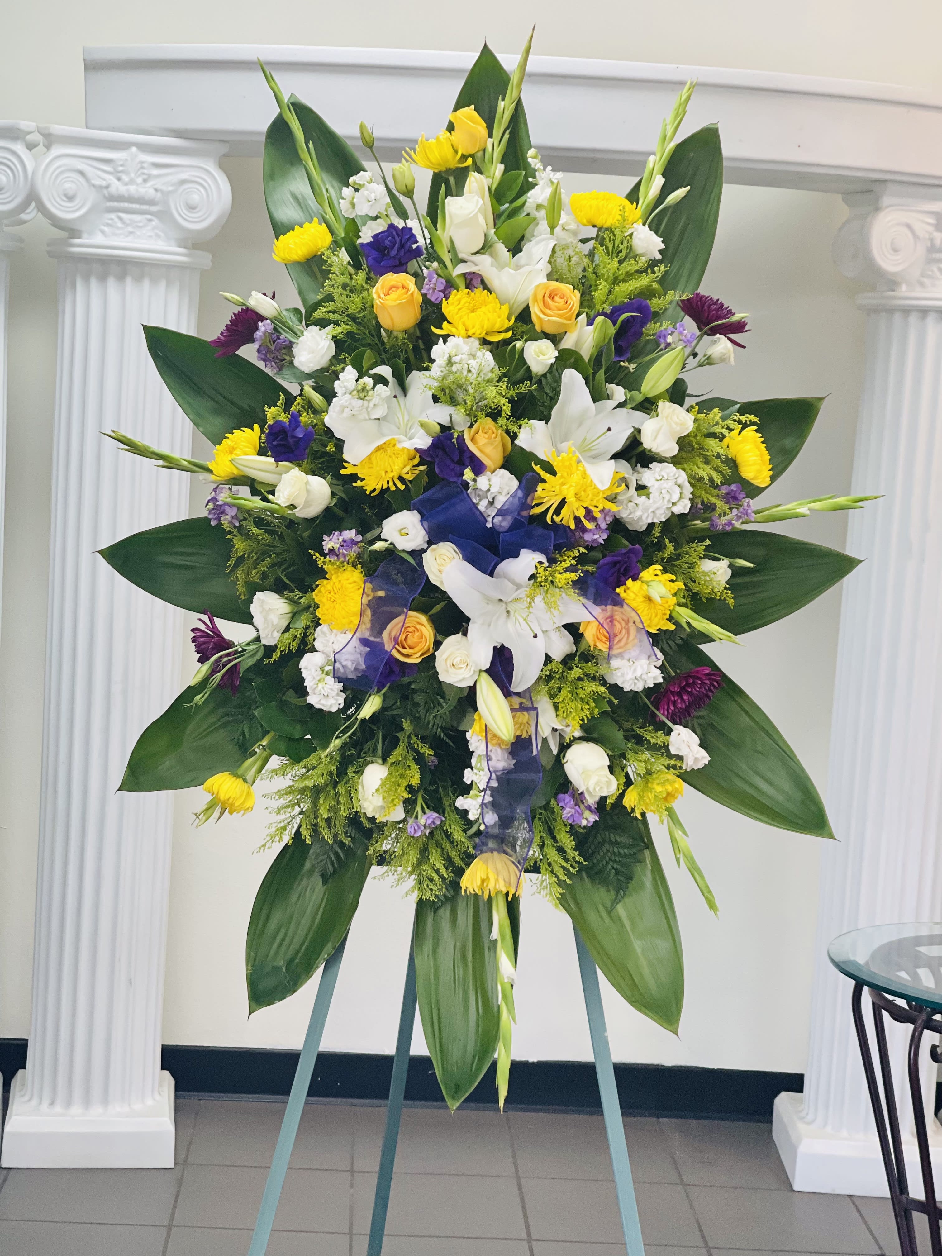 Tall standing spray of white lilies, yellow roses and purple blooms with large green leaves on an easel