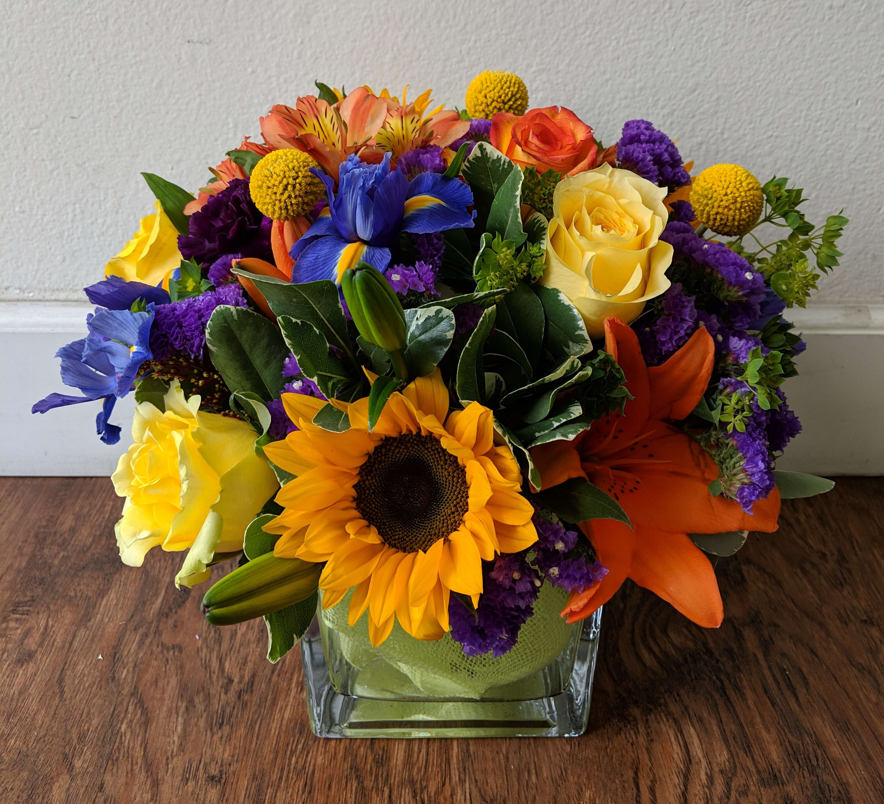 Colorful bouquet with sunflowers, roses, lilies, and purple filler in a square glass vase