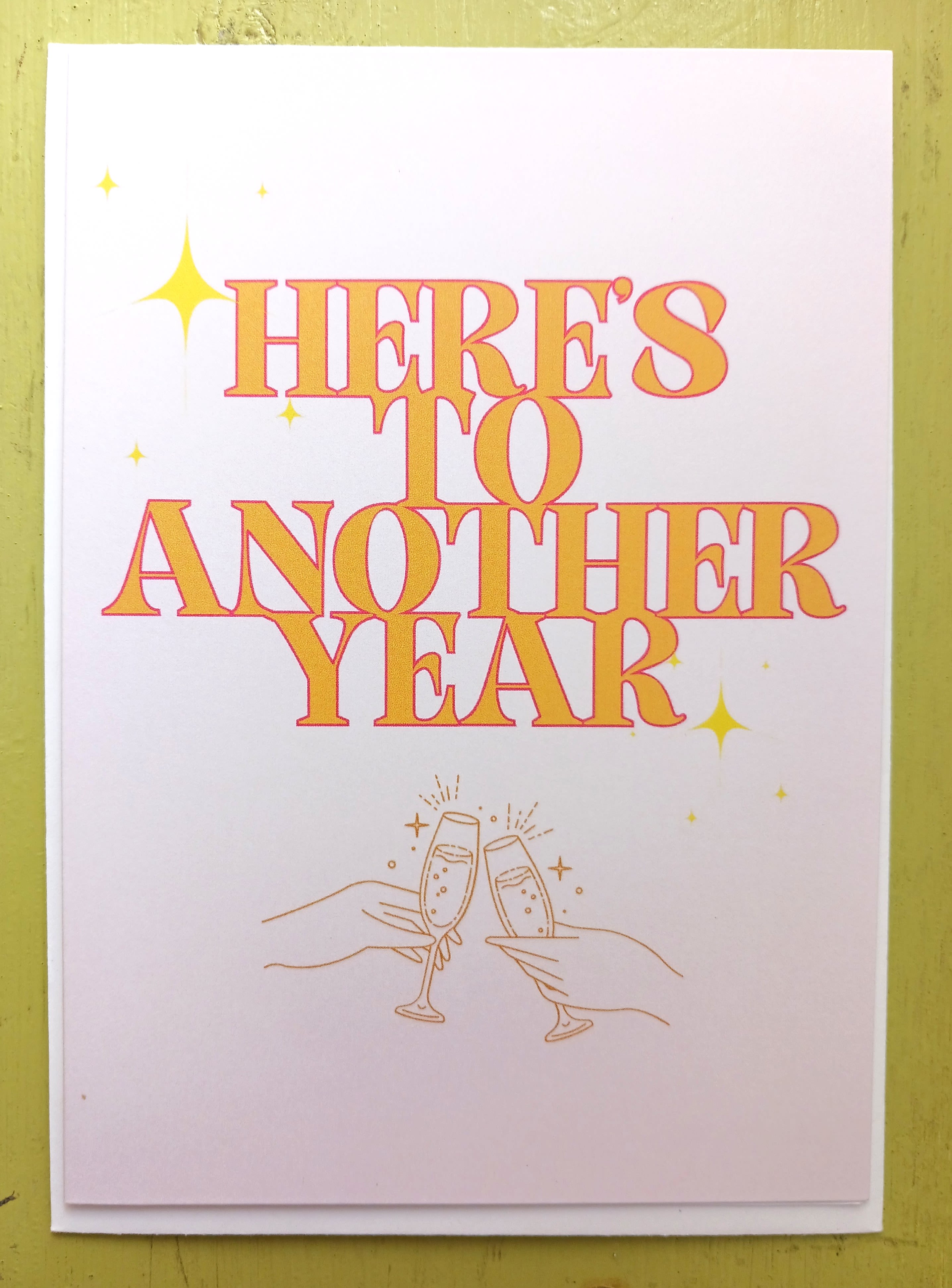 Greeting card with “Here’s to Another Year” text and hands clinking champagne glasses.