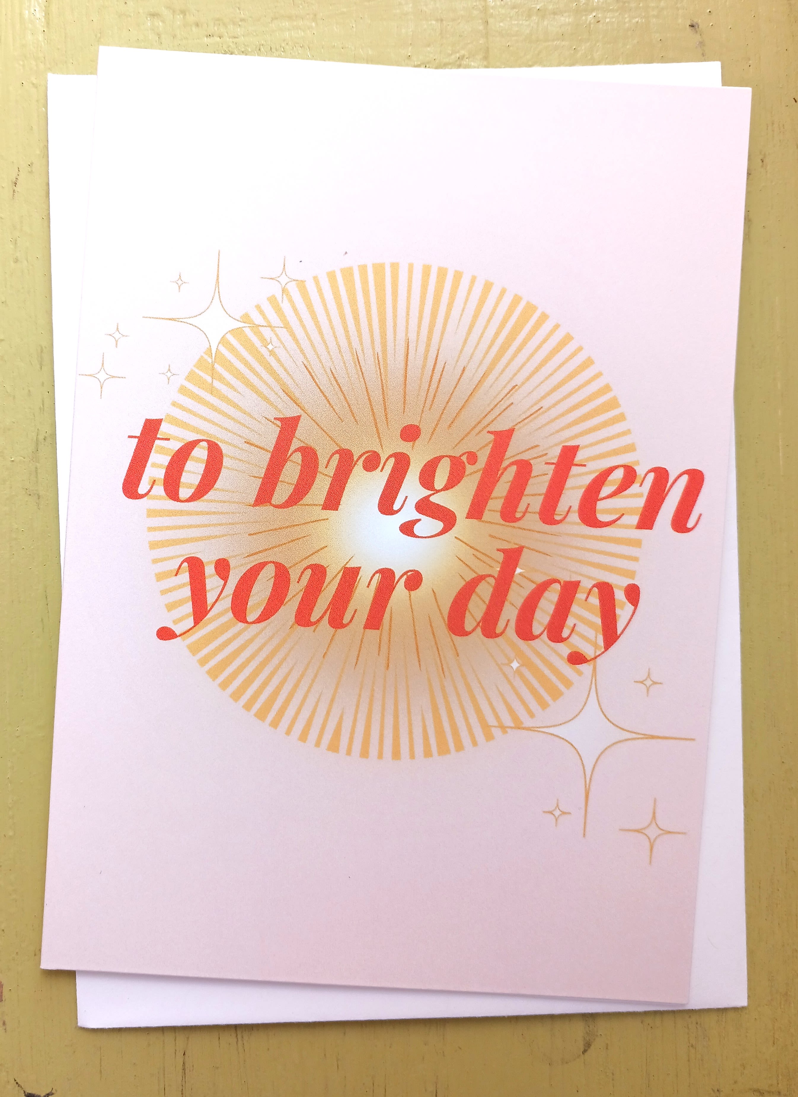 Folded pastel card with coral text 'to brighten your day' over a golden sunburst and small star accents.