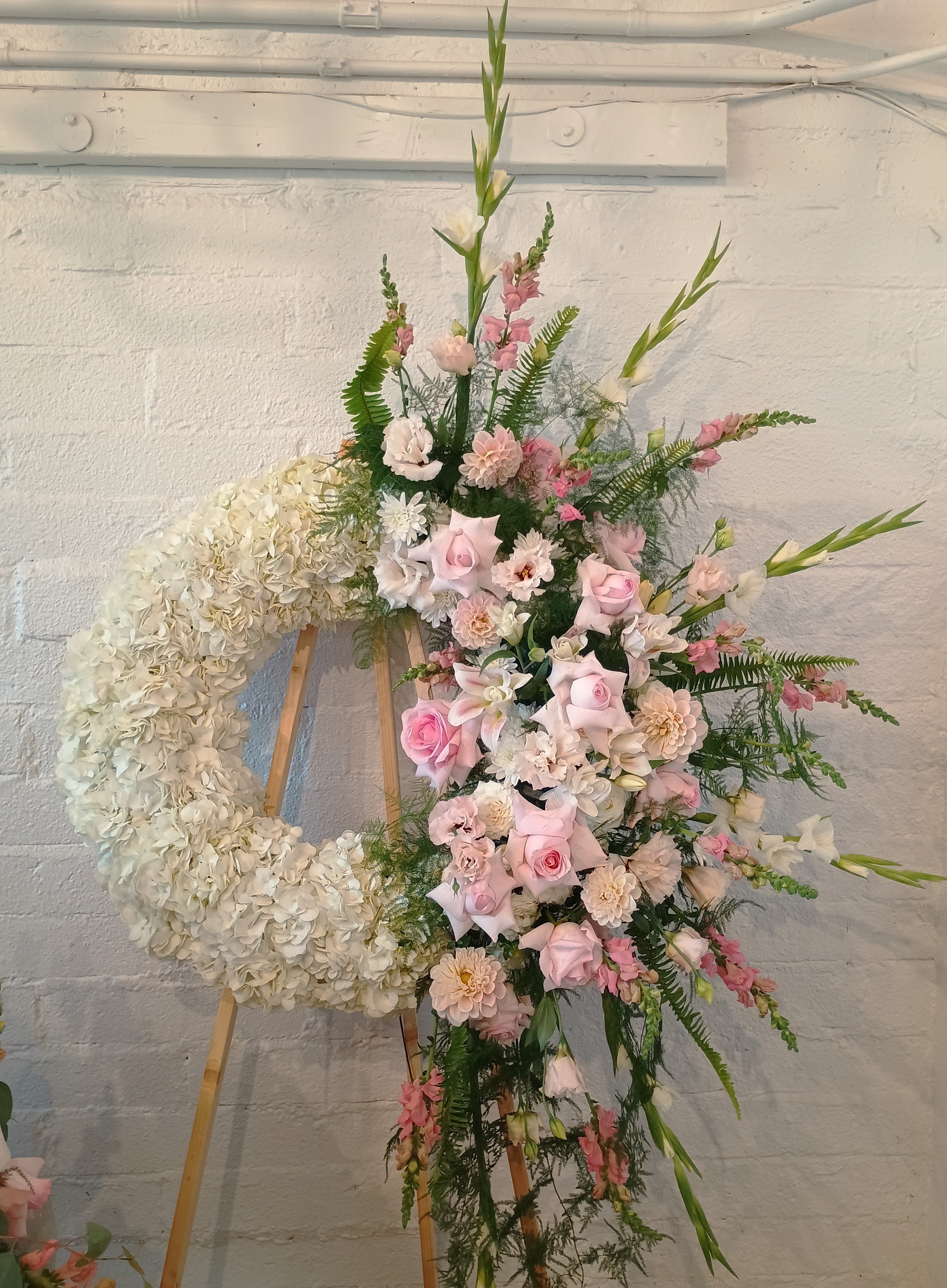 Pink and white floral funeral wreath on an easel beside a tall mixed arrangement