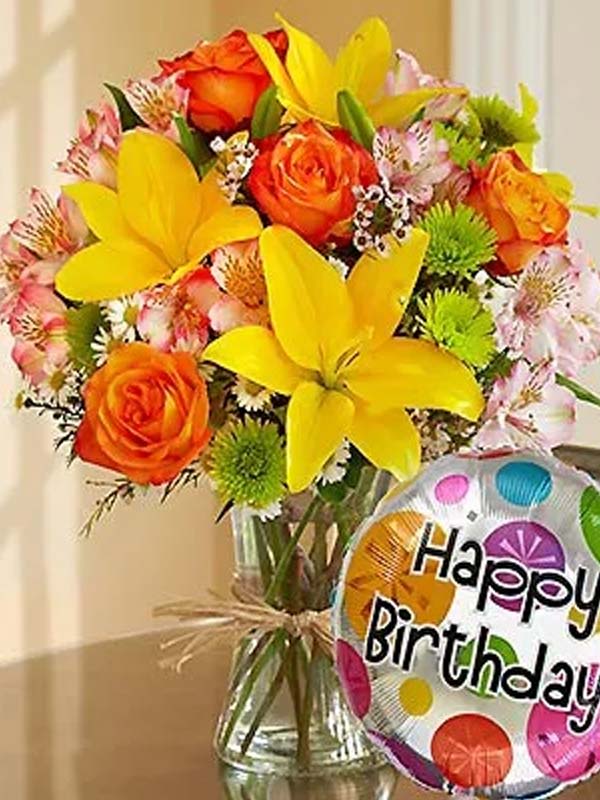 Bouquet of yellow lilies and orange roses in a glass vase with a birthday balloon