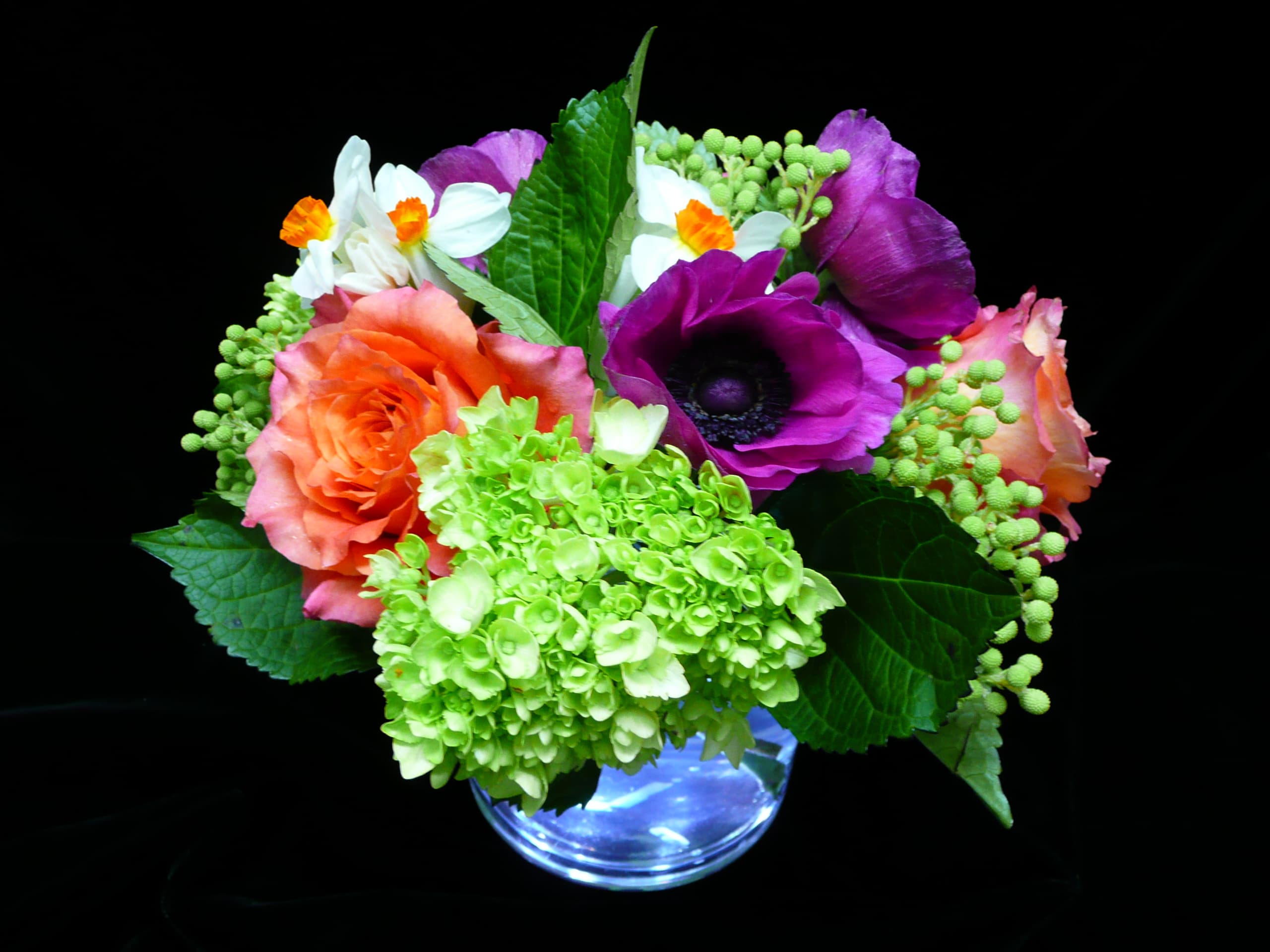 Bright mixed bouquet with orange roses, green hydrangea, purple anemone, and white flowers in a clear glass vase.