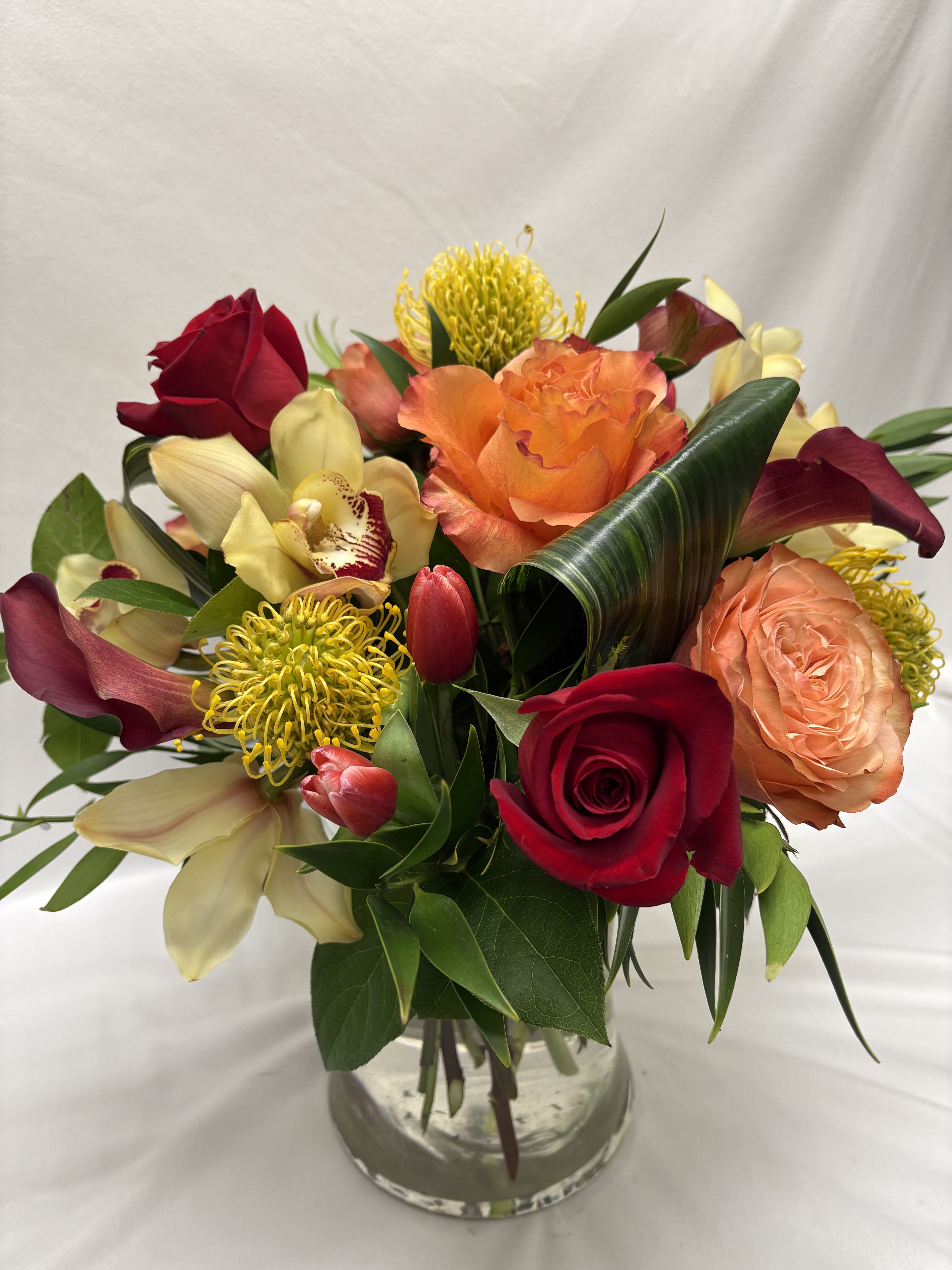 Vibrant arrangement of red and orange roses, yellow pincushion protea, orchids and tulips in a clear glass vase