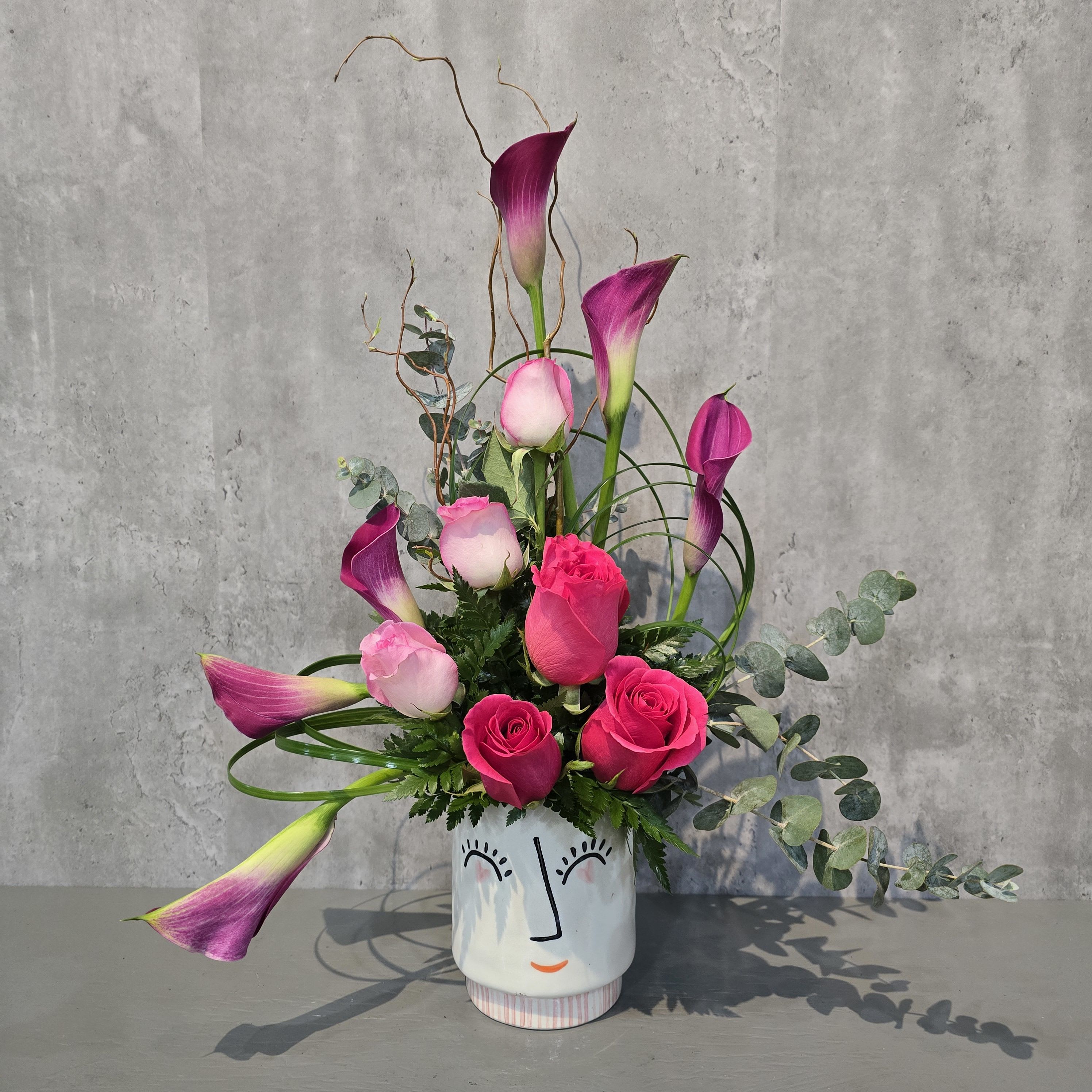 Blushing Goddess - **Blushing Goddess** is a breathtaking floral arrangement that embodies grace and femininity. Featuring lush pink roses and elegant calla lilies, this design blooms with soft romance and sophistication. Nestled in a ceramic vase adorned with a modern painted face, this arrangement feels like a work of art—both ethereal and timeless. Perfect for celebrating beauty, love, and strength, the **Blushing Goddess** makes a stunning gift for any occasion or a charming statement piece in any space.