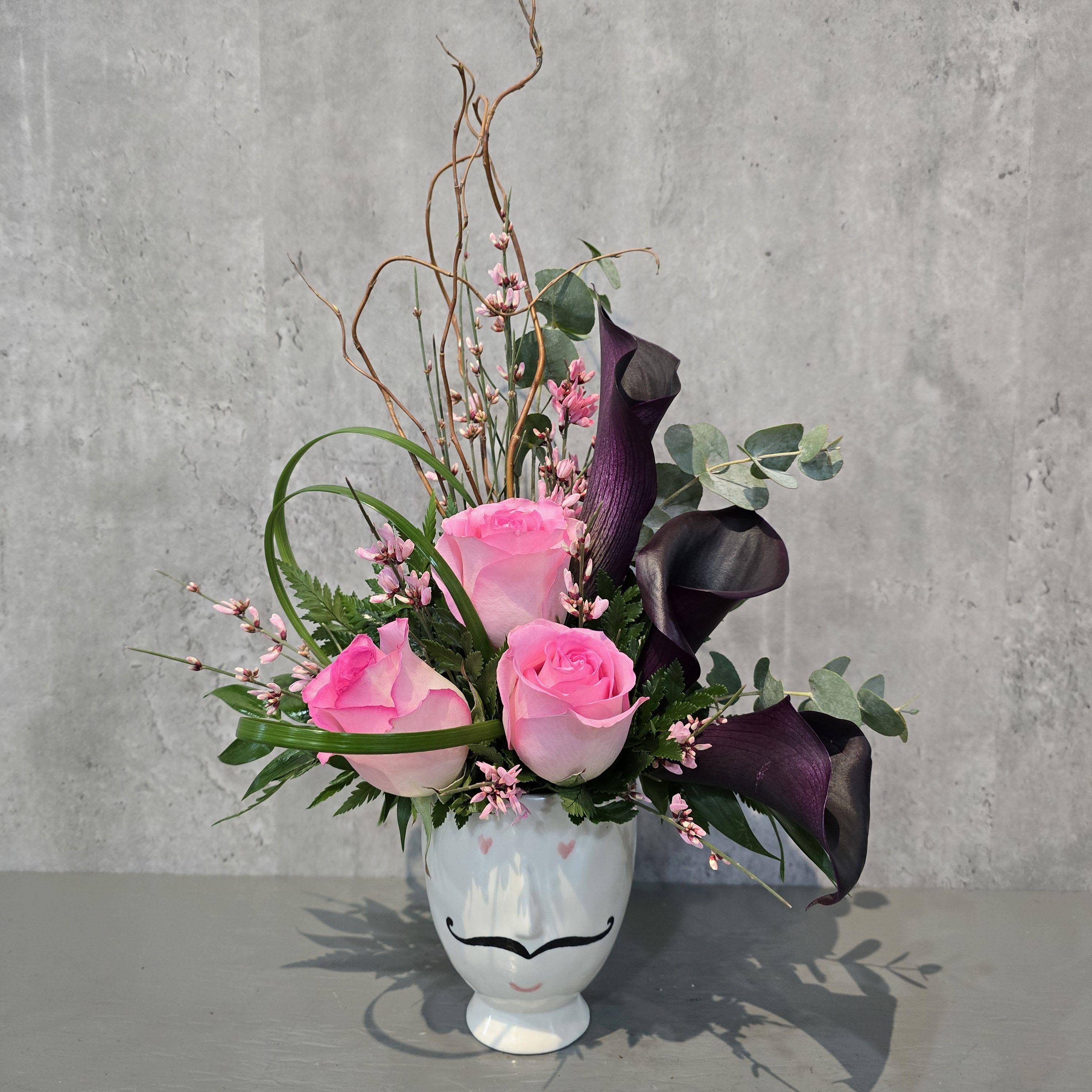 Monsieur L'Amour - Monsieur L'Amour is the perfect blend of Old-World charm and modern romance. This striking arrangement features lush pink roses and deep purple mini calla lilies, creating a dramatic yet elegant contrast. Housed in a small ceramic pot with a painted face reminiscent of a distinguished Italian gentleman, Monsieur L'Amour exudes sophistication and passion. Whether as a heartfelt gift or a statement piece, this floral creation brings a touch of European romance to any space.