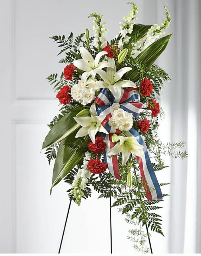 Standing floral spray with white lilies, red carnations, and ribbon on an easel