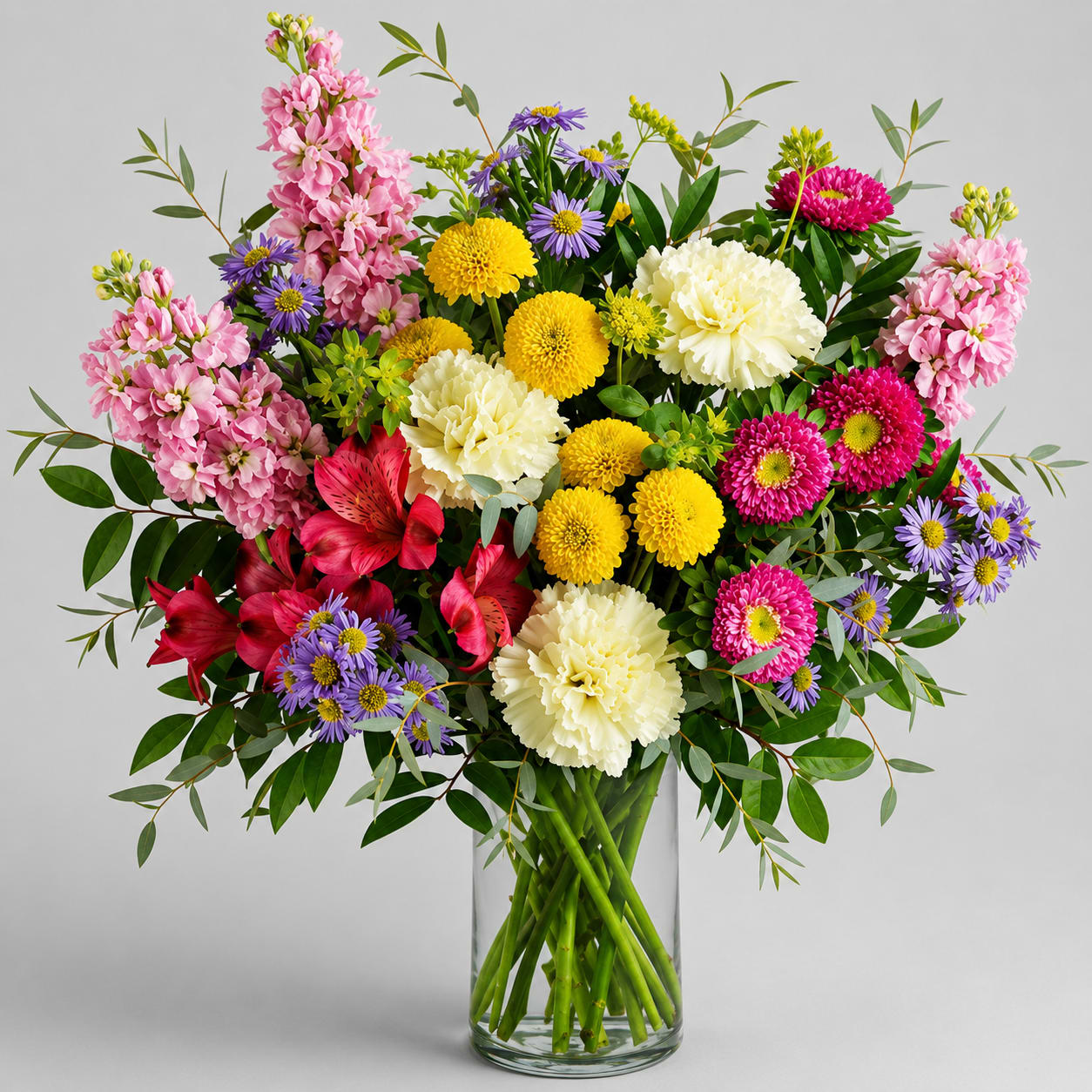 Nature's Beauty - A bright, garden-style arrangement that reads like a fresh-picked bouquet-perfect as a cheerful get well gift, housewarming welcome, or spring/summer celebration. This mixed-color flower arrangement features pink stock and carnations, magenta alstroemeria, yellow button chrysanthemums, white blooms, and purple daisy-like asters that together create a lively yet soft palette your recipient will notice the moment it arrives.  Flowers are arranged loose and abundant in a clear glass vase so stems and water show through for a natural, airy look; greenery branches outward to give the design a relaxed, garden feel. The combination balances long-lasting, sturdy blooms (chrysanthemums and carnations) with fragrant, romantic stock and delicate asters-an everyday bouquet that still feels special for Women's Day, Best Friends Day, Grandparents' Day, or Admin Professionals' Week. Simple care-fresh water and a clean vase-keeps the mix looking its best.
