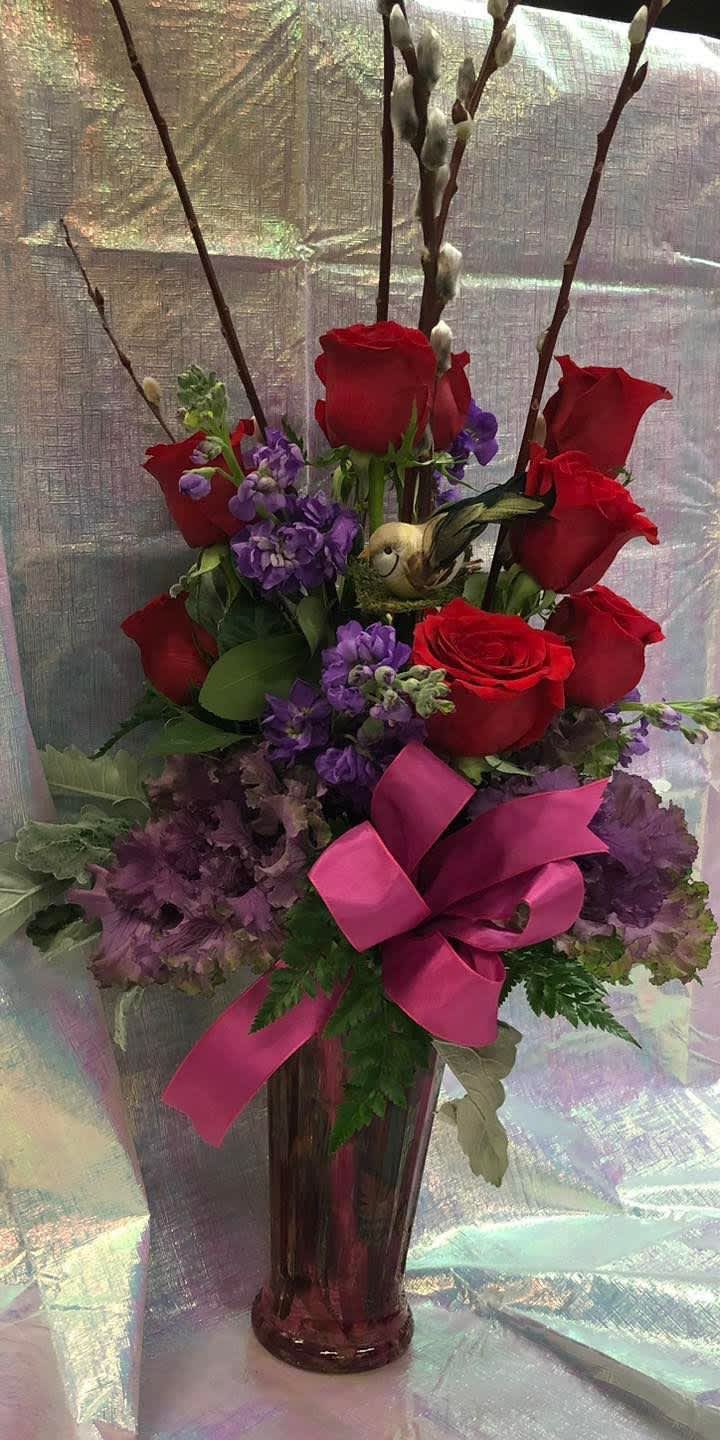 Red roses and purple flowers in a tall glass vase with a pink ribbon