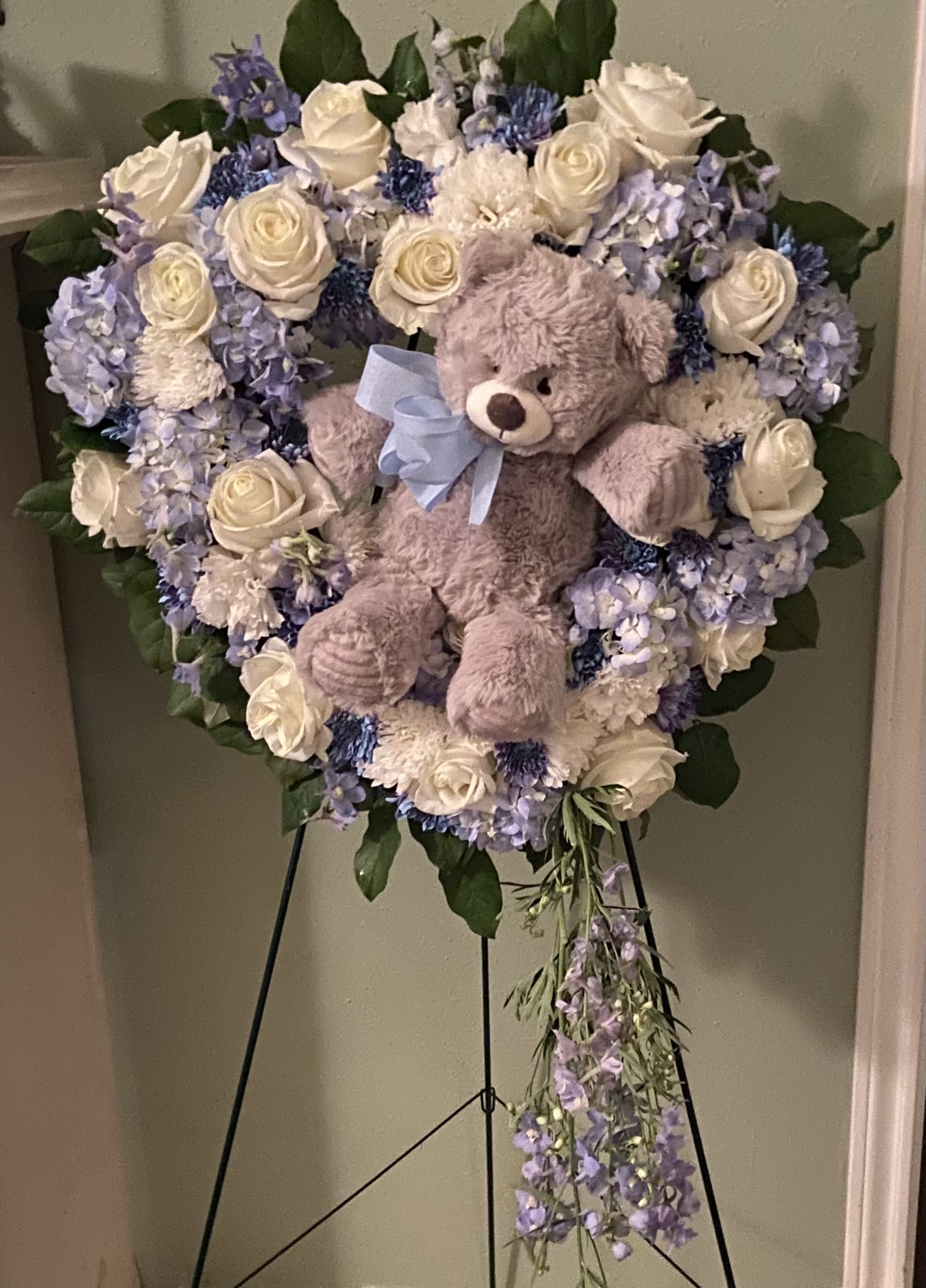Heart-shaped floral wreath with a teddy bear and blue ribbon on an easel