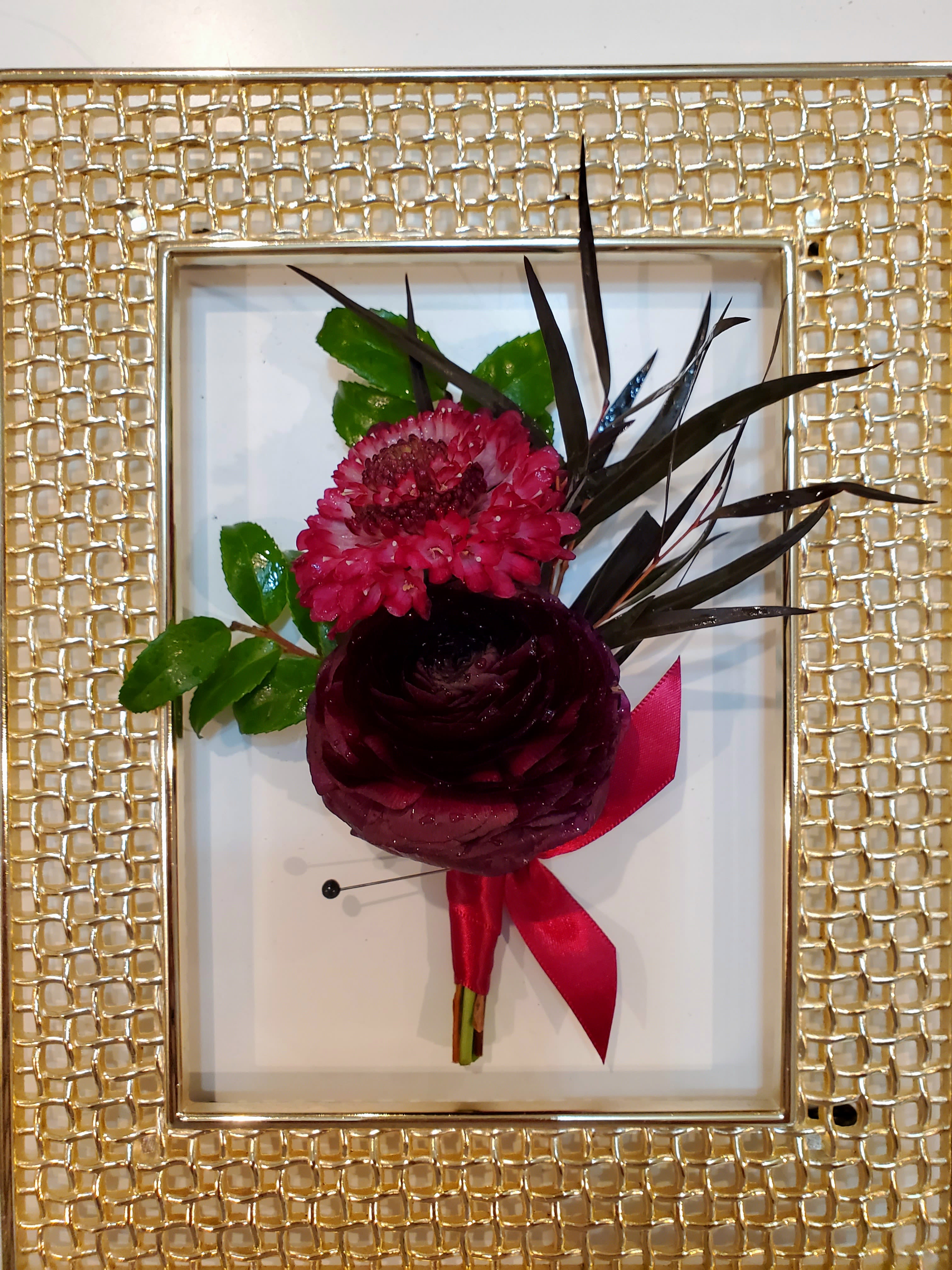 Small floral boutonniere with dark red blooms and a pink ribbon in a gold frame