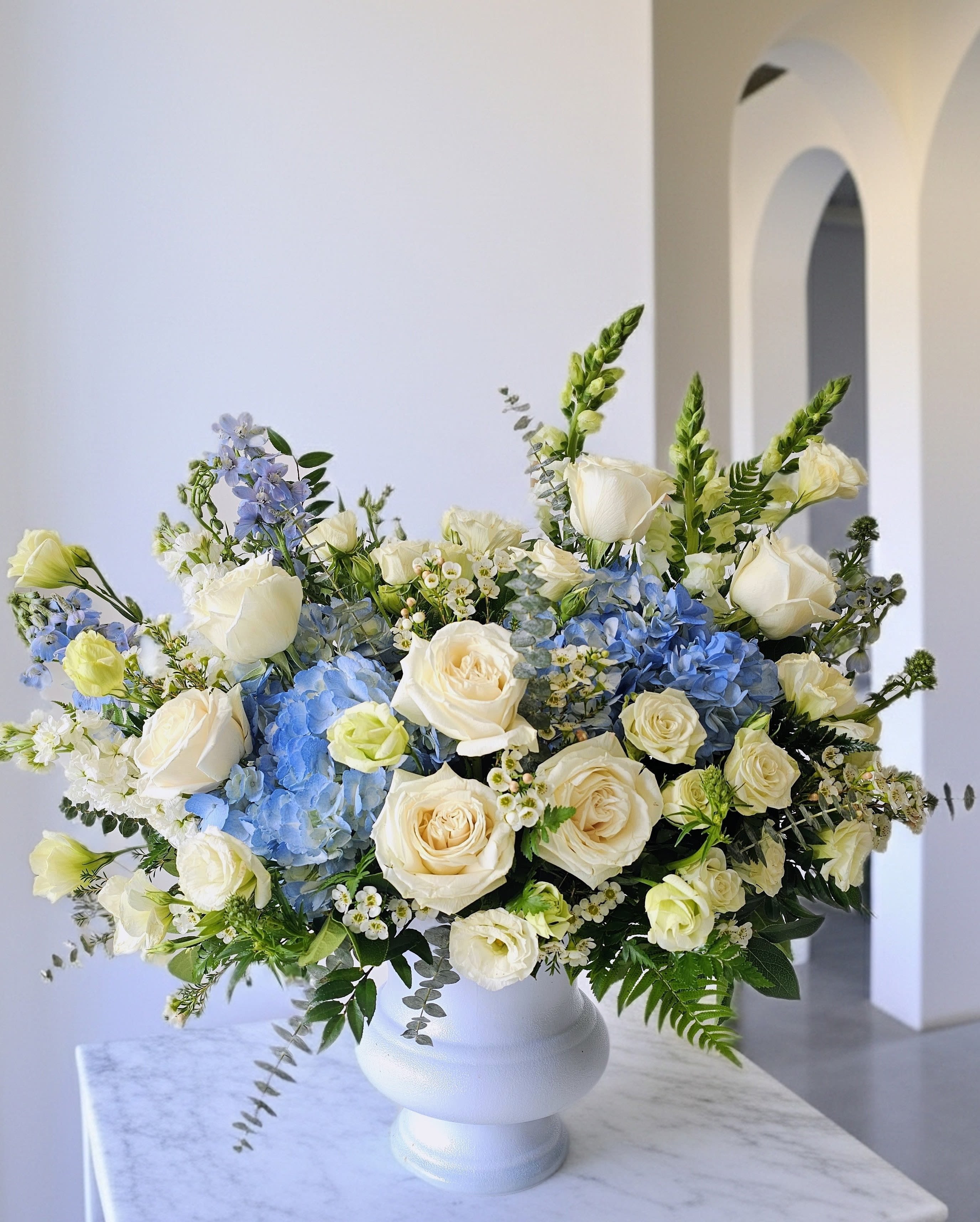 Blessed From Above Urn Basket - Cream-white roses and pale blue hydrangeas create a soft, garden-fresh impression in this funeral and wedding flower arrangement, perfect as a sympathy tribute or an elegant ceremony centerpiece. The airy mix of lisianthus, delphinium spikes, snapdragons, stock and delicate filler blooms gives a refined, peaceful presence that will help the person you're honoring feel remembered and seen.  Arranged asymmetrically for a natural, flowing silhouette, stems and green foliage reach outward from a clean white ceramic vase for a lush, full presentation. The palette of white, blue and soft greens keeps the look serene and versatile for memorial needs or wedding décor. Sized to make a thoughtful statement without overwhelming a space, it arrives ready-to-display; water daily and keep in cool, indirect light to extend freshness.