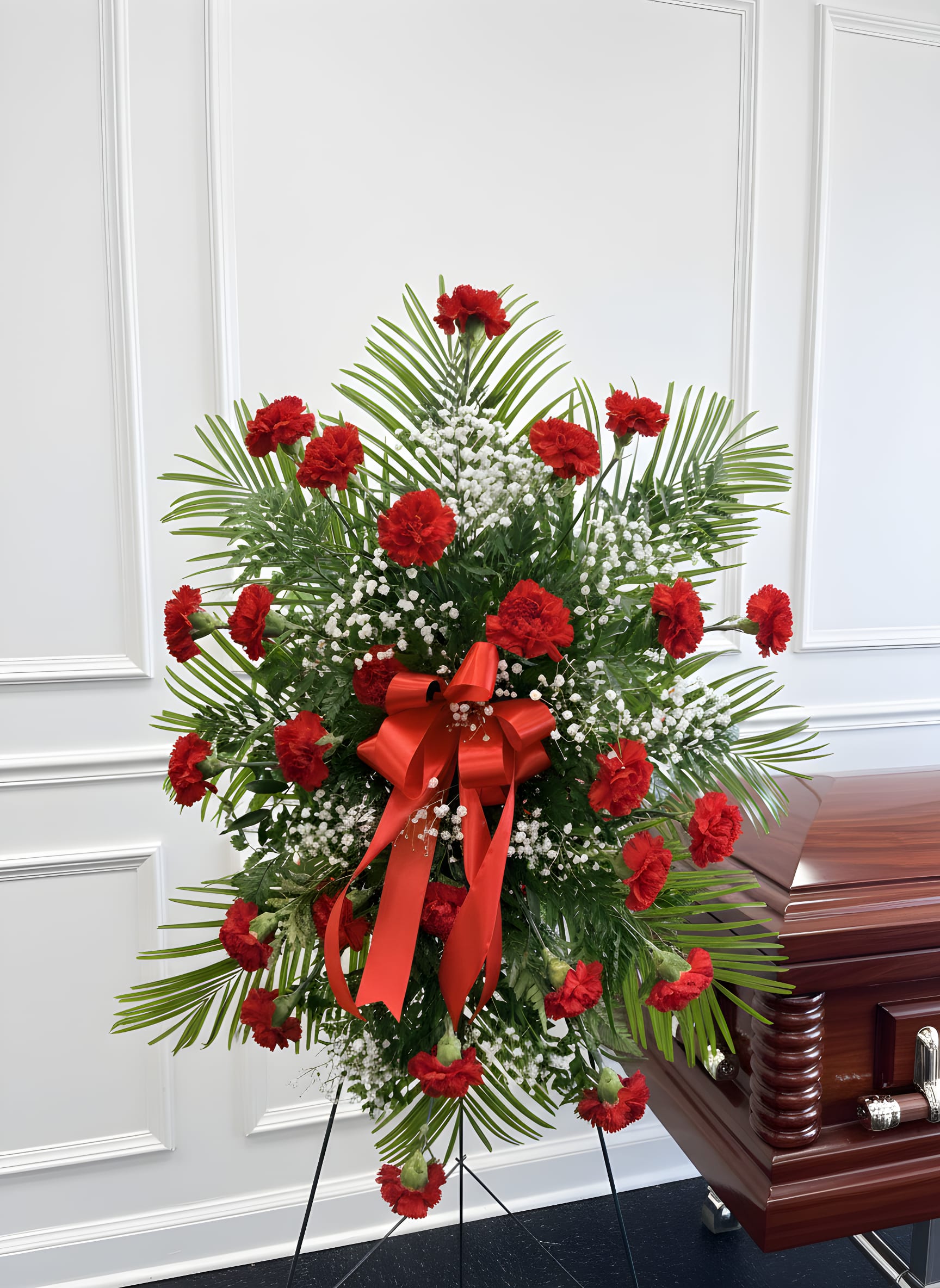Forever in Our Hearts Red Carnation Standing Spray - Honor a cherished life with the timeless beauty of our Forever in Our Hearts Red Carnation Standing Spray. This elegant funeral standing spray is handcrafted with vibrant red carnations, delicate white baby's breath, lush premium greenery, and finished with a coordinating satin ribbon. Displayed on a sturdy easel, this sympathy arrangement offers a meaningful expression of love, respect, and remembrance.  Red carnations symbolize deep love, admiration, and enduring affection, making this standing spray a beautiful tribute for funeral services, memorial ceremonies, celebrations of life, or graveside services. Carefully designed by our professional florists using the freshest flowers available, this arrangement brings comfort to family and friends during life's most difficult moments.  Perfect For:  Funeral Services Memorial Services Celebration of Life Ceremonies Sympathy Tributes Graveside Services