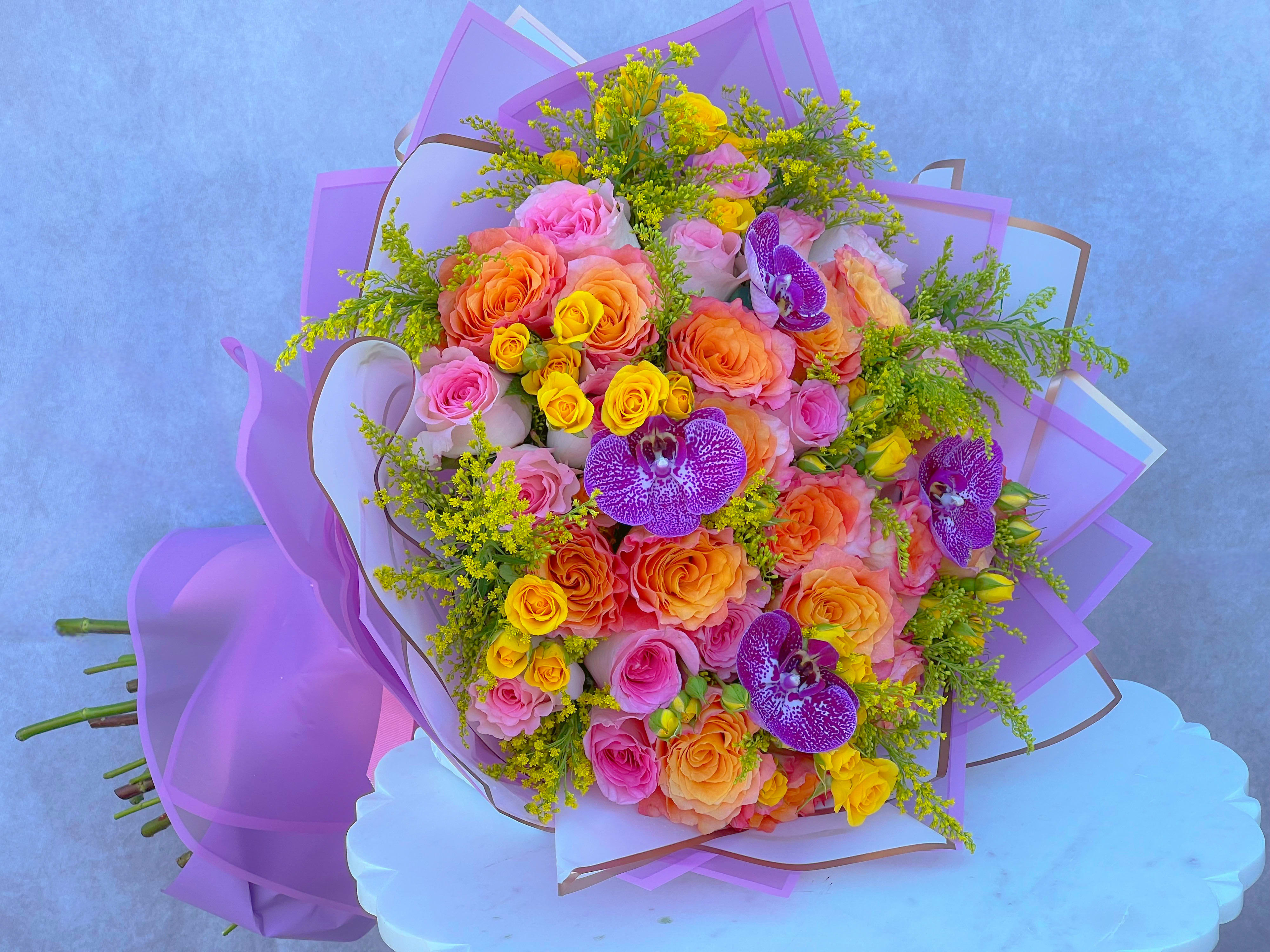 Large bouquet of pink, orange, and yellow roses with purple orchids wrapped in lavender paper.