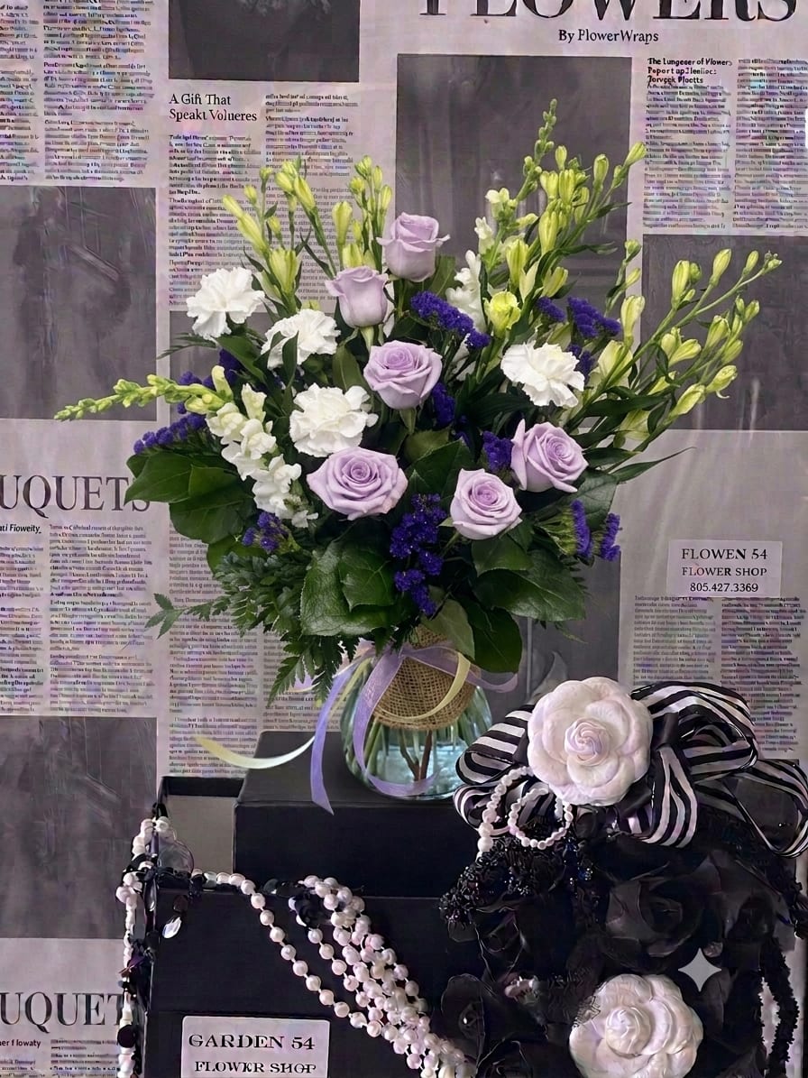 Lavender roses and white flowers arranged in a glass vase with ribbons.