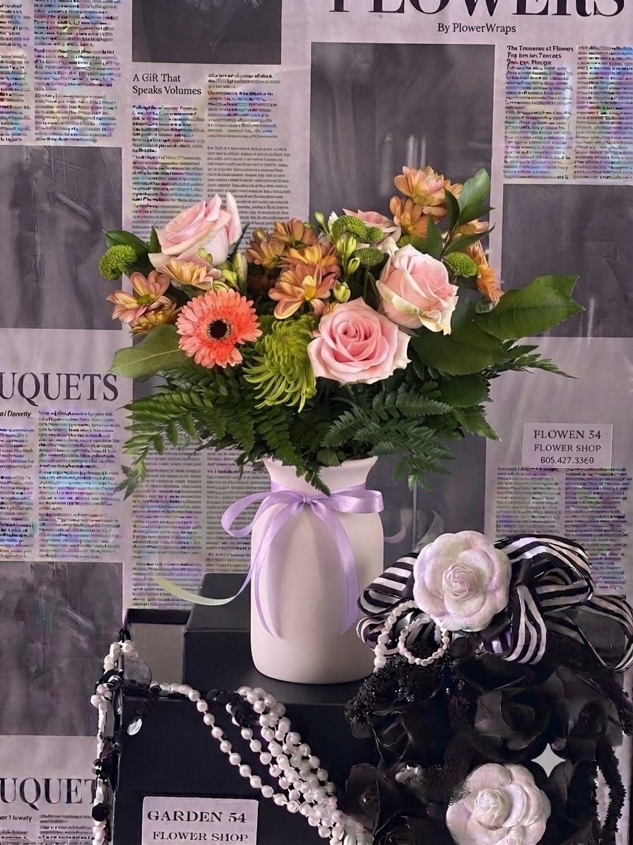 Pink and peach mixed flowers arranged in a white vase with a lavender ribbon.