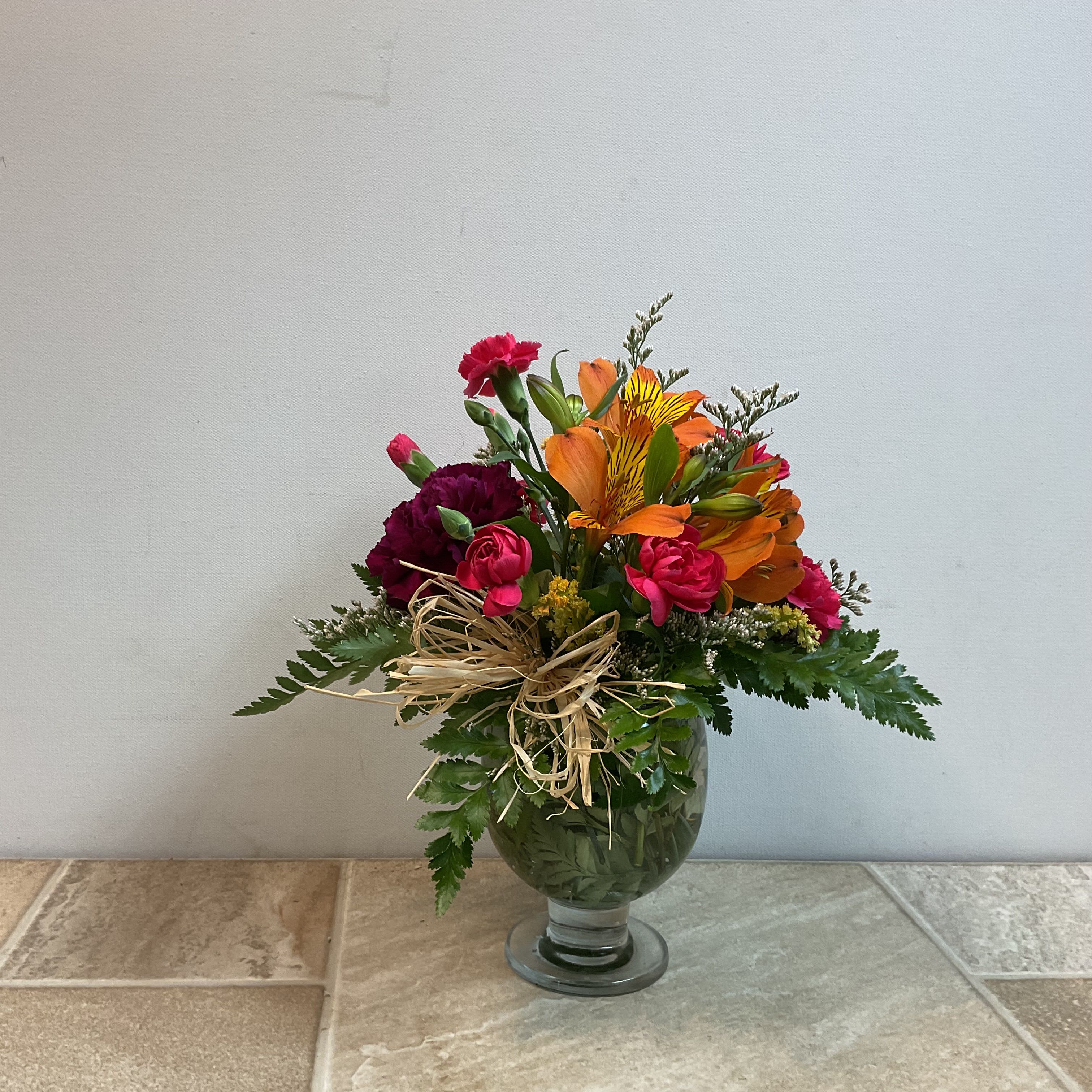 Itty Bitty Bouquet  - A lively, compact mixed flower arrangement that makes someone's day - perfect as a birthday bouquet, thank-you gift, or just-because surprise. This flower arrangement features bright orange alstroemeria, cheerful pink mini carnations and purple carnations in a rounded, slightly asymmetrical shape so the recipient feels instantly noticed and delighted.  Tightly gathered in a low clear glass vase and finished with a rustic raffia bow, the Itty Bitty Bouquet is designed for tabletops and small spaces without losing floral impact. The mixed-color palette of orange, pink, and purple offers energetic contrast against dense greenery; its size is intentionally petite for easy placement and immediate enjoyment. It measures 10” wide x 12” tall.   Primary flowers: lily, carnation