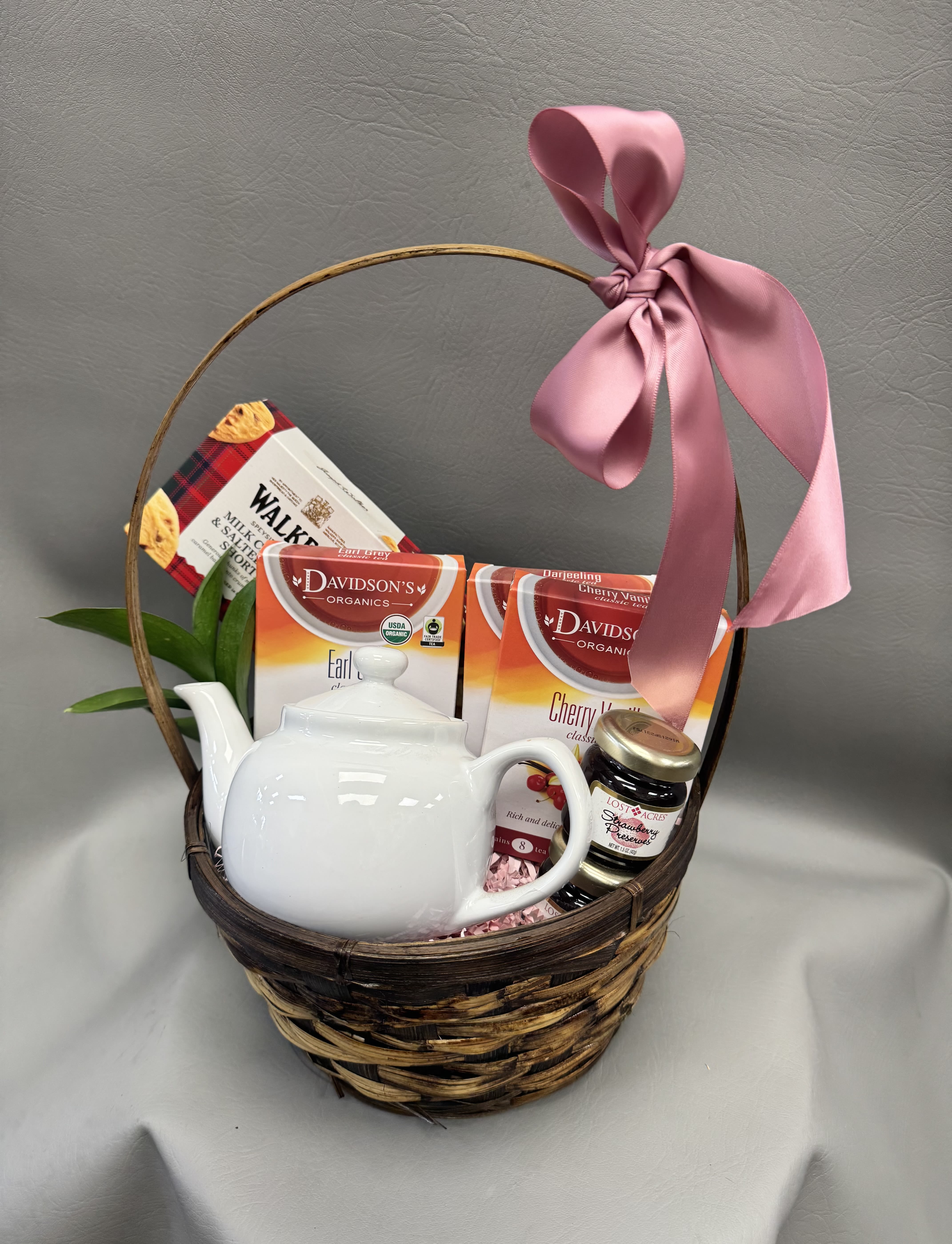 Tea Basket - White teapot and assorted teas in a round wicker basket with a pink satin bow and jar of strawberry preserves-soft, cozy tea-time gift perfect for thank-yous and casual celebrations.