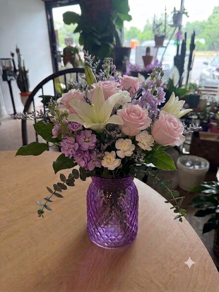Amethyst dreams - Pale pink roses and white lilies mingle with lavender blooms and cream carnations to create a soft, romantic flower arrangement perfect as a birthday bouquet, anniversary gift, or thoughtful thank-you. The composition's pastel palette - pink, white, purple and touches of green - reads fresh and springlike, giving the person you're thinking of an immediate sense of calm and delight.  Arranged compactly in a purple cut-glass vase, the mix of textures - the roses' velvety petals, the lilies' graceful trumpets, the ruffled carnations and delicate filler blooms - feels both lush and refined. The purple glass adds a striking color contrast that turns the vase itself into a keepsake decorative accent, so the recipient can enjoy it beyond the flowers.  This is the kind of arrangement that makes someone feel truly seen: gentle, elegant, and perfectly suited for everyday gifting or a special small celebration. The compact design fits tabletops and desks; refresh water and trim stems every few days to extend bloom life.