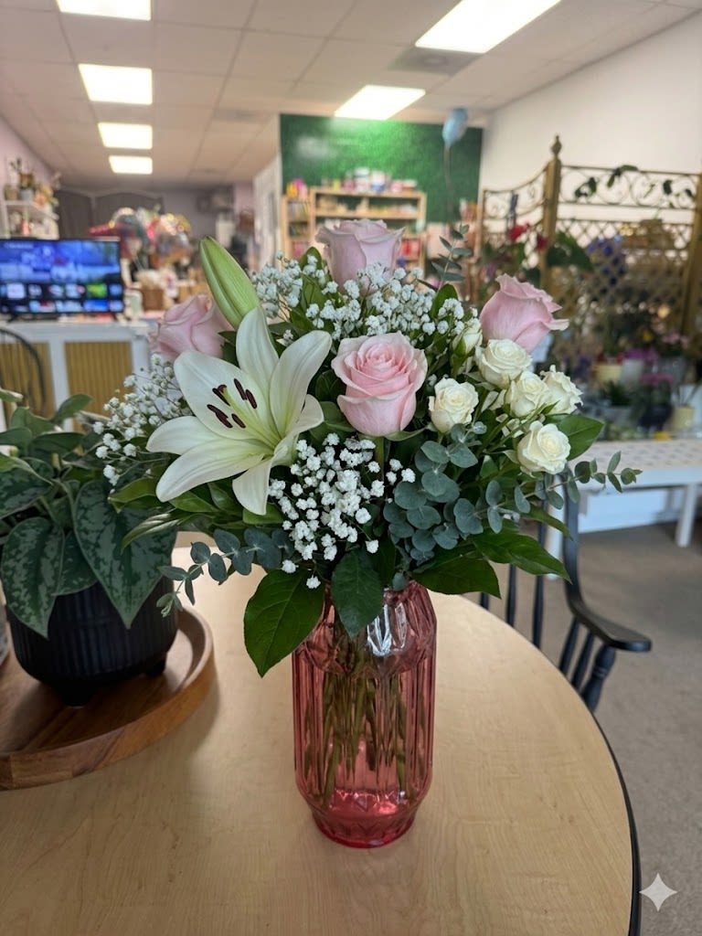 Pink Promise - A soft cluster of pale pink and white roses crowned by a single white lily creates a gentle, romantic statement-perfect as a birthday bouquet, anniversary roses, or a thoughtful "thinking of you" arrangement. The upright shape and airy mix of blooms give the person you're sending it to an immediate sense of calm and care, the kind of bouquet that reads as both intimate and put-together.  This flower arrangement features pale pink roses, white roses, a prominent white lily, delicate white filler blooms, and rounded eucalyptus-like green foliage arranged in a tall pink glass vase. The pink glass adds a whisper of color at the base while keeping the overall palette clean and classic; mixed textures-velvety rose petals, sleek lily blooms, and soft greenery-create a polished, balanced look that photographs beautifully and holds its shape.  Ideal for celebrating special moments or offering comfort, Pink Promise suits recipients who appreciate timeless, elegant florals. The tall vase gives it presence on a dining table or entry console; order with any available size options to match the occasion, and enjoy simple care-fresh water in the vase and a cool location will keep the bouquet looking its best.