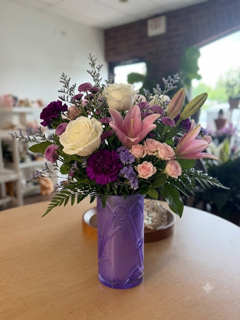 Lavender Twilight - Soft pink lilies and creamy white roses curl above clusters of blush spray roses and deeper purple accents in this Lavender Twilight flower arrangement-an elegant birthday bouquet or anniversary roses alternative that feels both romantic and fresh. The rounded composition and coordinated violet tones give the person you're thinking of a sense of being cherished, while the deeper purple blooms add a lively contrast that keeps the look modern and spirited.  This arrangement features lilies, roses, carnations, chrysanthemums and complementary filler blooms arranged in a compact, rounded shape and set in a striking purple glass vase that anchors the mixed color palette. Textures range from the lilies' silky petals to the velvety roses and the small, ruffled carnations, creating visual depth in shades of purple, pink, and white. The vase's bold color acts as a stylish accent-no additional container needed.  Perfect for birthdays, anniversaries, or a thoughtful thank-you, Lavender Twilight reads as a polished everyday bouquet that makes the recipient feel truly seen. Designed to arrive ready-to-display, it brings a long-lasting presence to a bedside table or dining centerpiece; enjoy changing water every couple of days and trimming stems for best freshness.