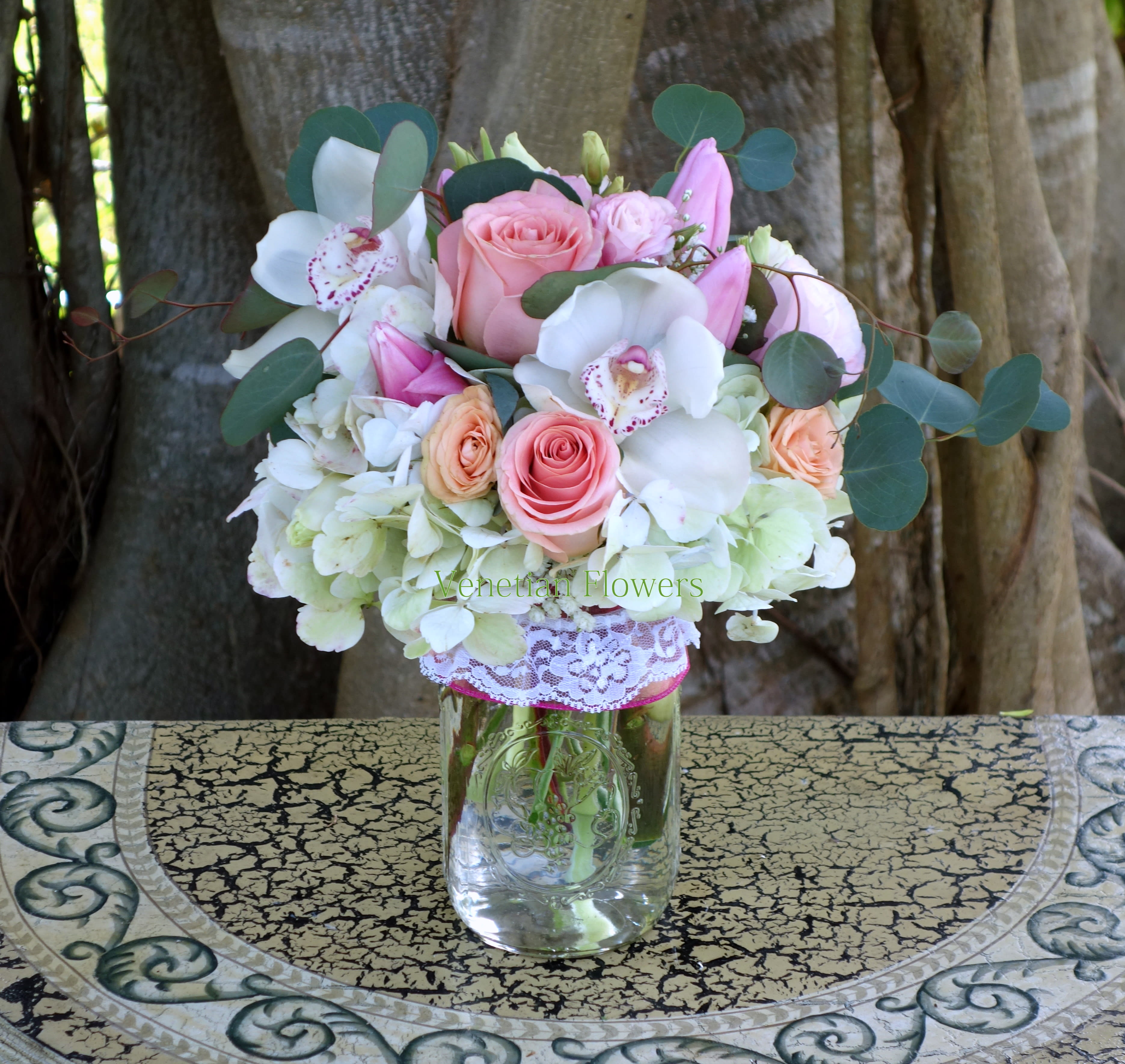 Mason jar arrangement with pink and peach roses, white orchids, hydrangeas, and pink lilies.