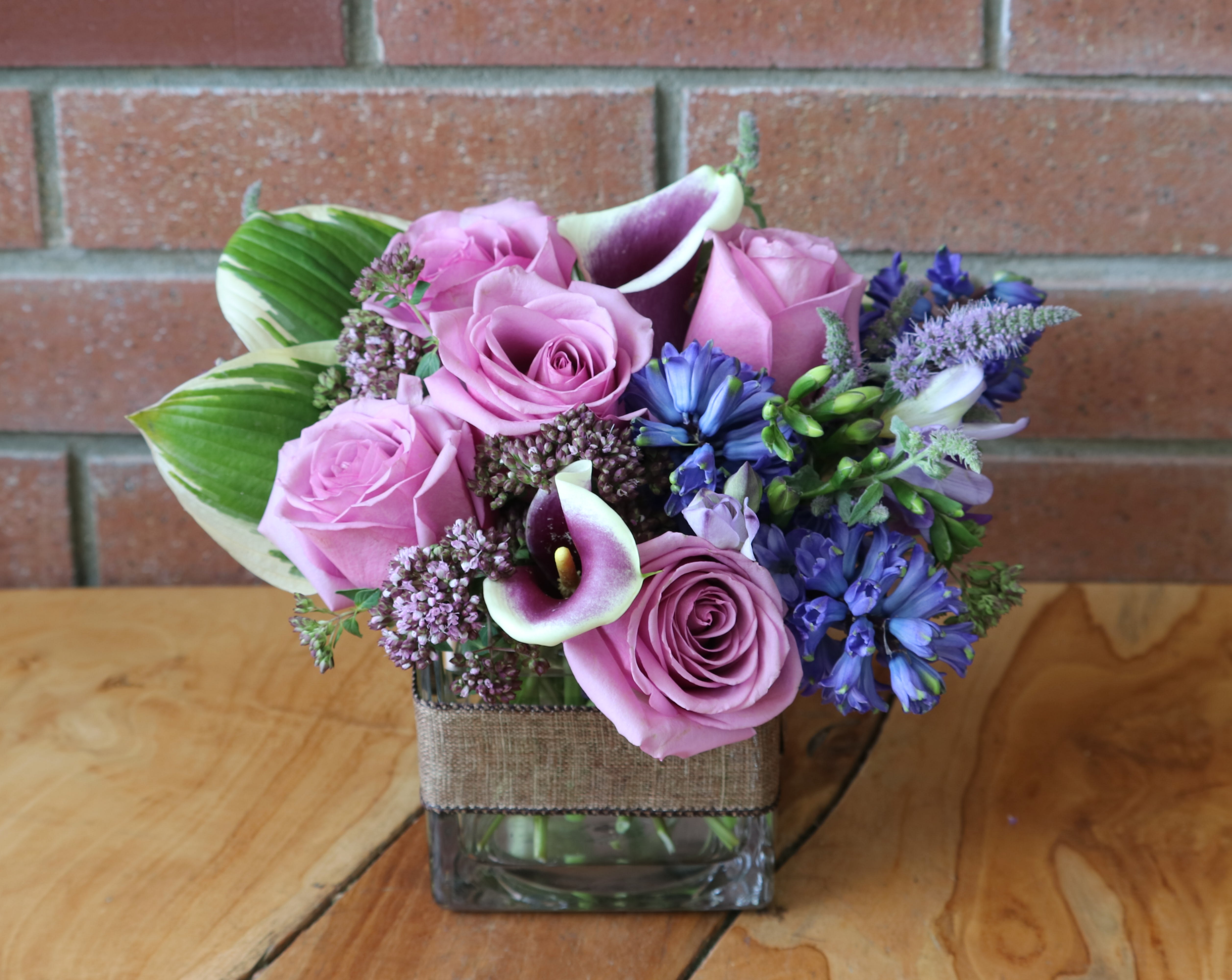 Short arrangement of pink roses, purple calla lilies, and blue flowers in a square glass vase