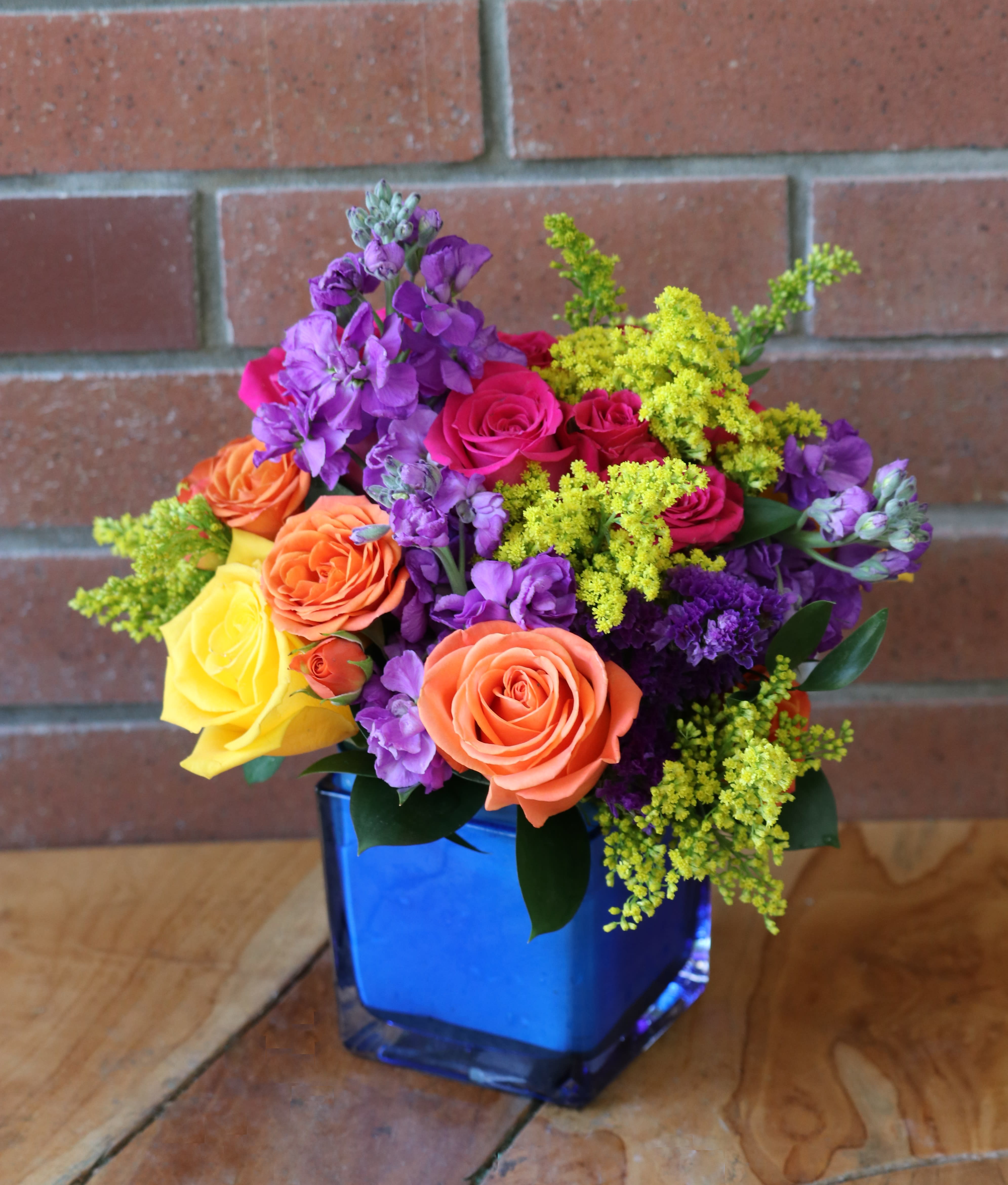 Bright mixed bouquet of orange and yellow roses with purple flowers in a blue glass cube vase