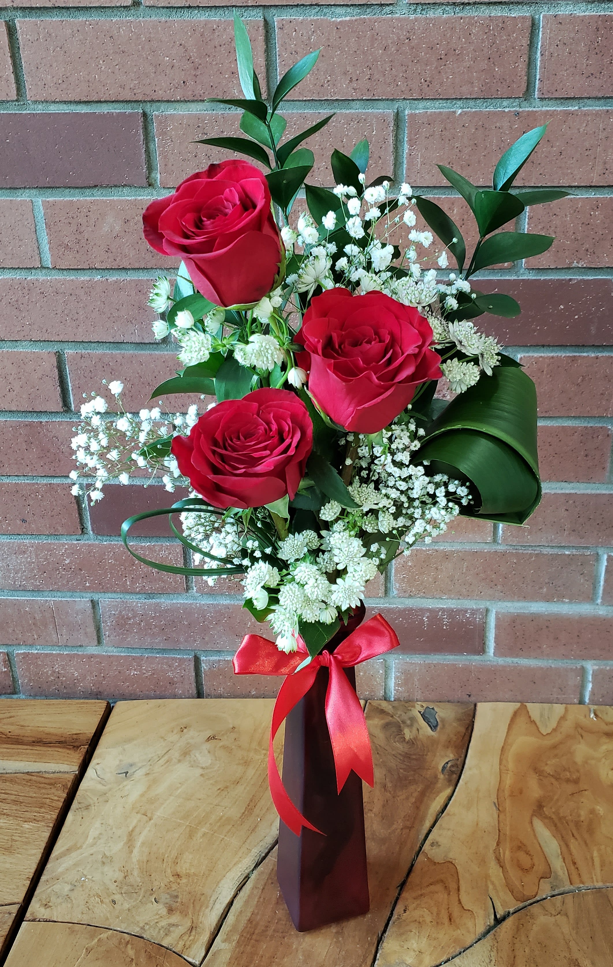 Tall red vase with three red roses and white filler flowers tied with a red ribbon