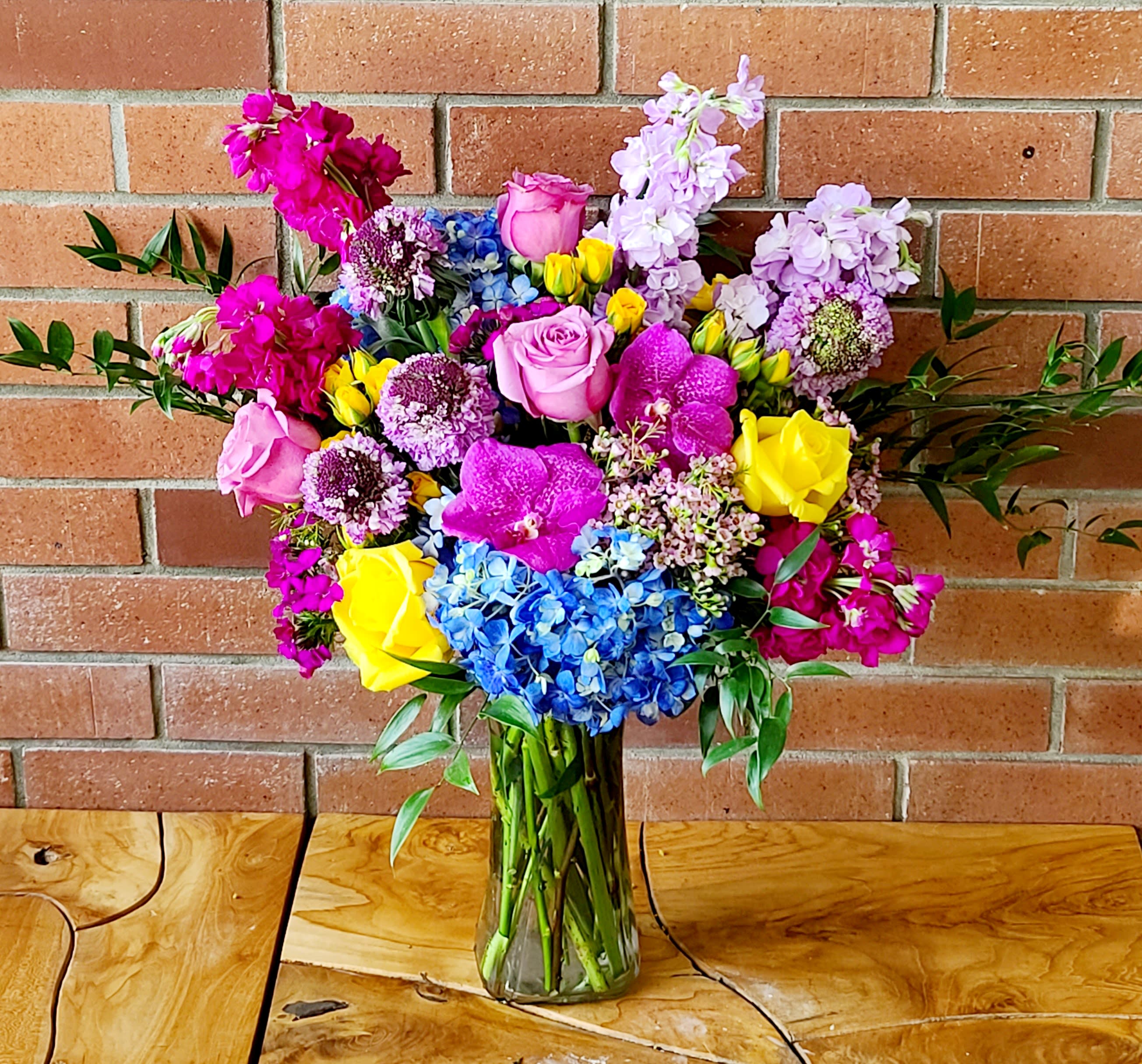 Colorful bouquet of roses, orchids, hydrangeas, and purple blooms in a glass vase