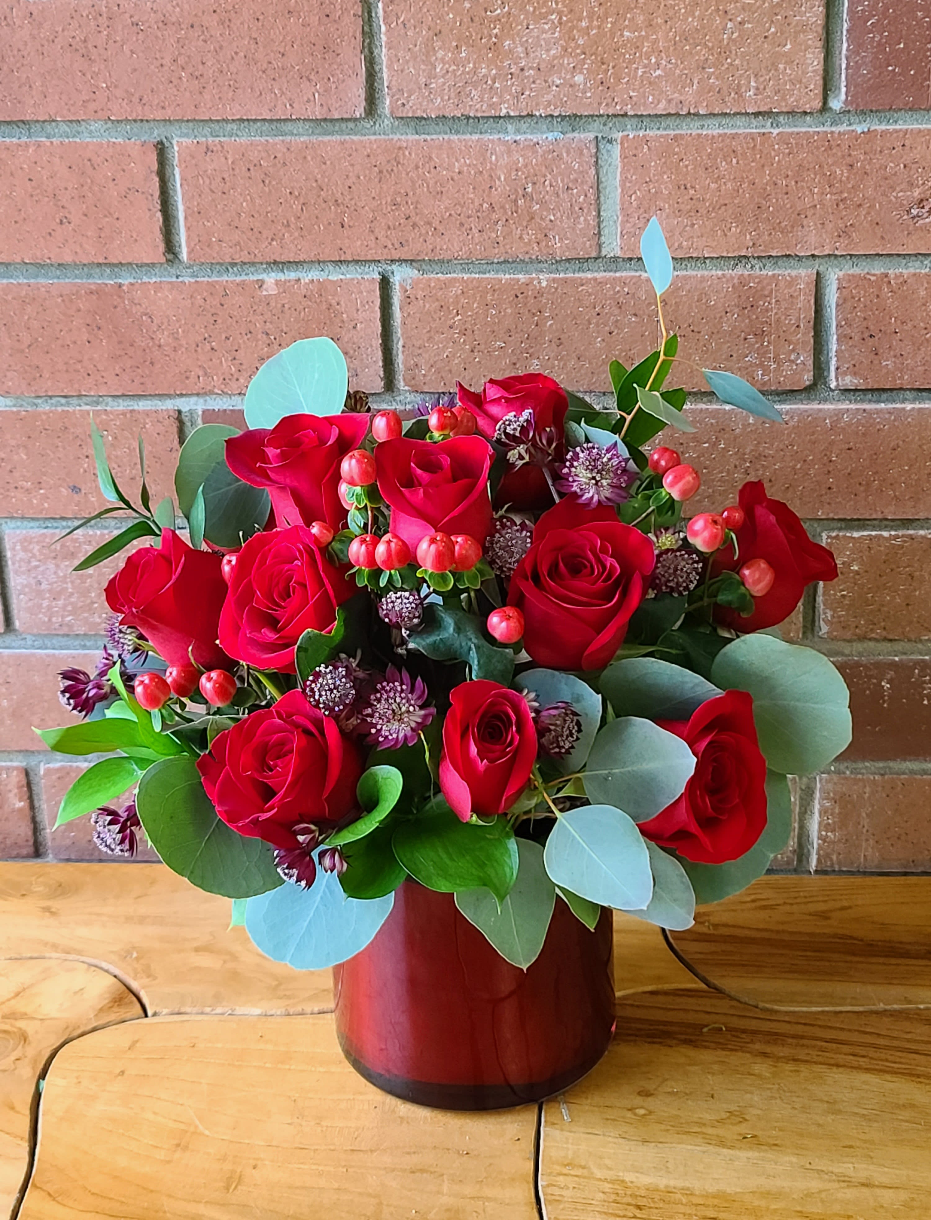 Red roses arranged in a red vase with eucalyptus and small accent flowers