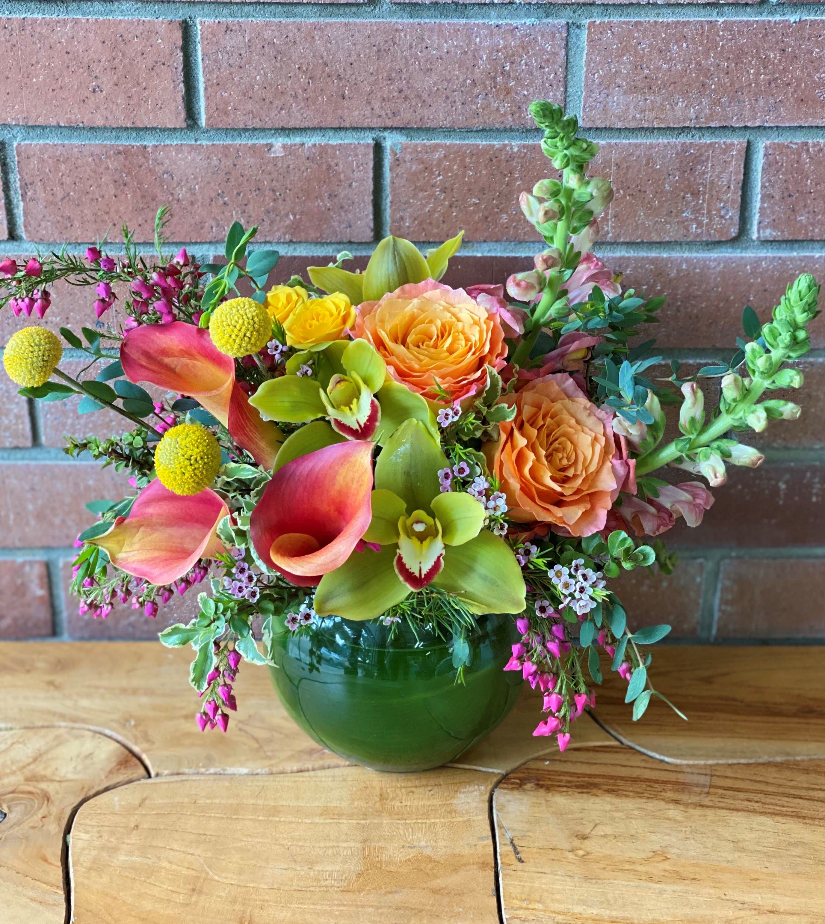 Low arrangement of orange roses, pink calla lilies, and green orchids in a round green glass vase