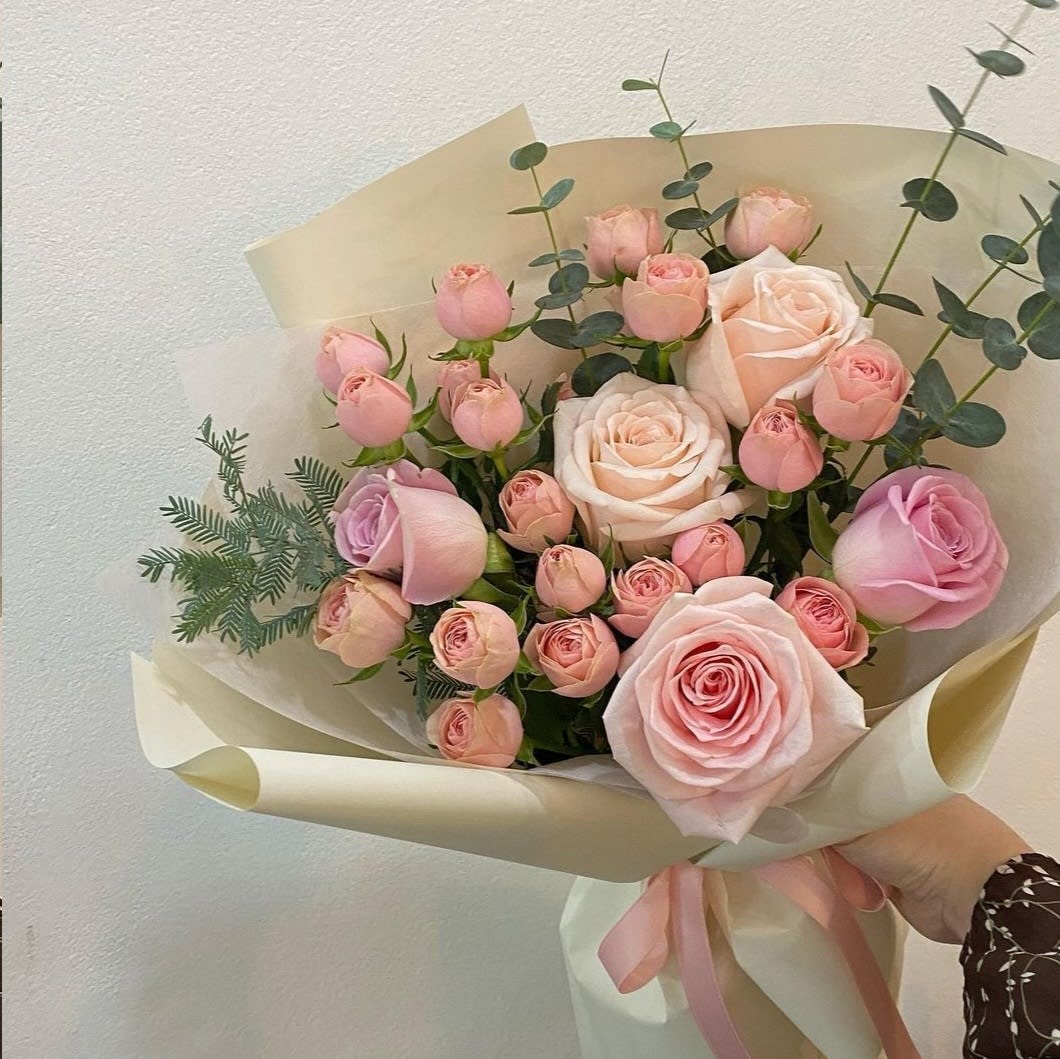 Hand-tied bouquet of light pink and peach roses and rosebuds wrapped in cream paper with a pink ribbon